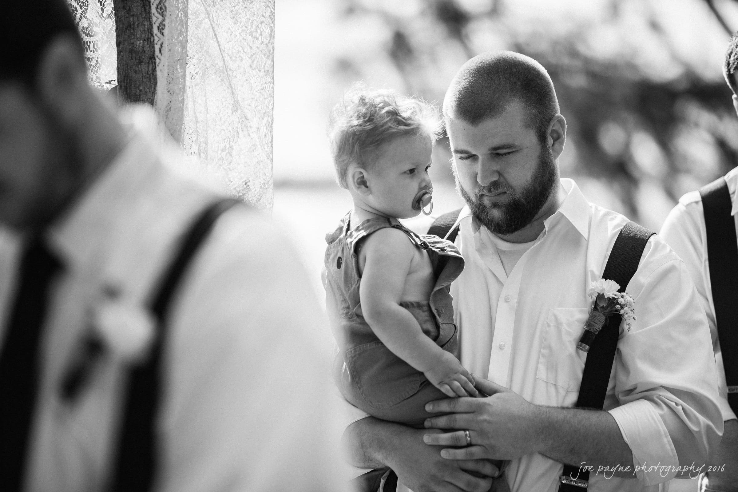 new bern wedding photographer - jessica & brinson A bearded man in a white shirt and suspenders lovingly holds a small child with a pacifier. The scene is in black and white, capturing a candid, tender moment during a formal event.
