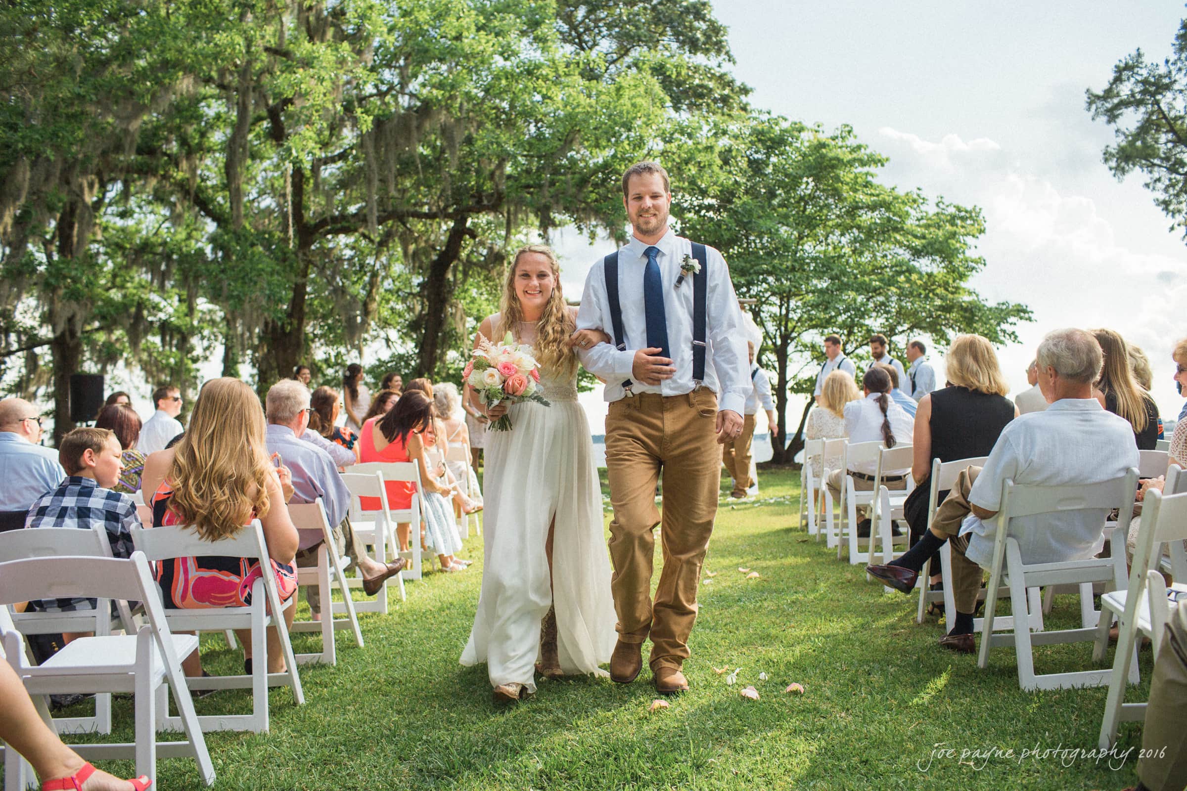 new bern wedding photographer - jessica & brinson A couple, smiling and dressed in wedding attire, walk down an outdoor aisle lined with seated guests on white chairs. The background features lush green trees and a sunny sky.