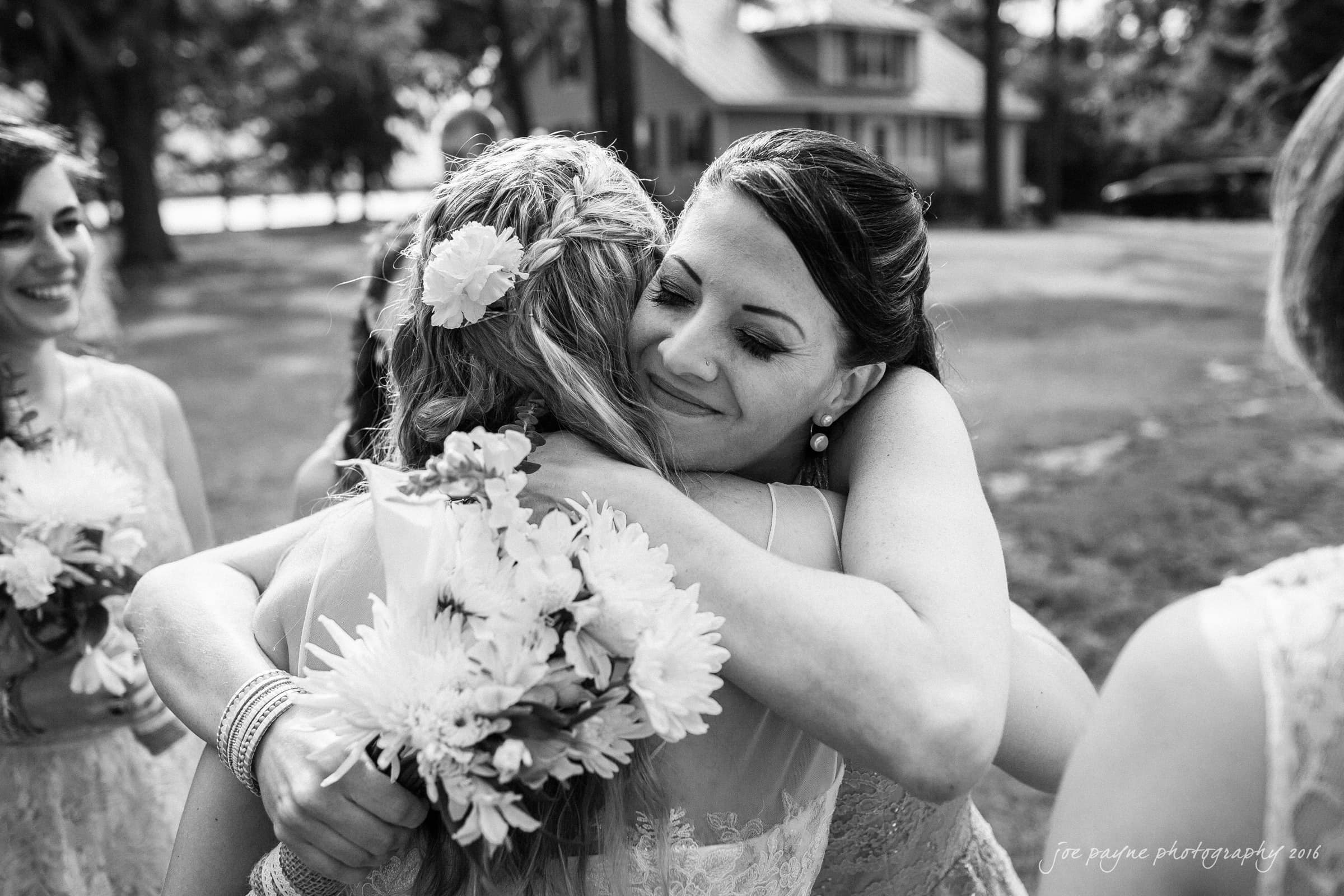 new bern wedding photographer - jessica & brinson A black and white photo of two women embracing outdoors, surrounded by blurred trees and a house in the background. One woman, smiling, holds a bouquet of flowers. Others stand nearby, also holding flowers.