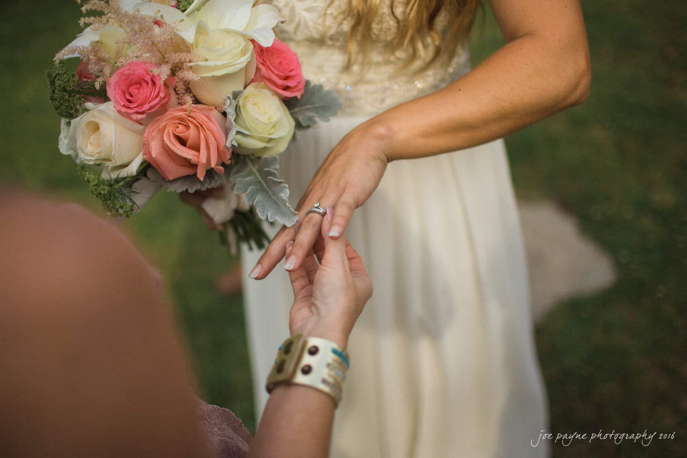 new bern wedding photographer - jessica & brinson A woman in a white dress, holding a bouquet of white and pink flowers, extends her hand to another person. They are both focused on an engagement ring on her finger. The scene takes place outdoors with green grass in the background.