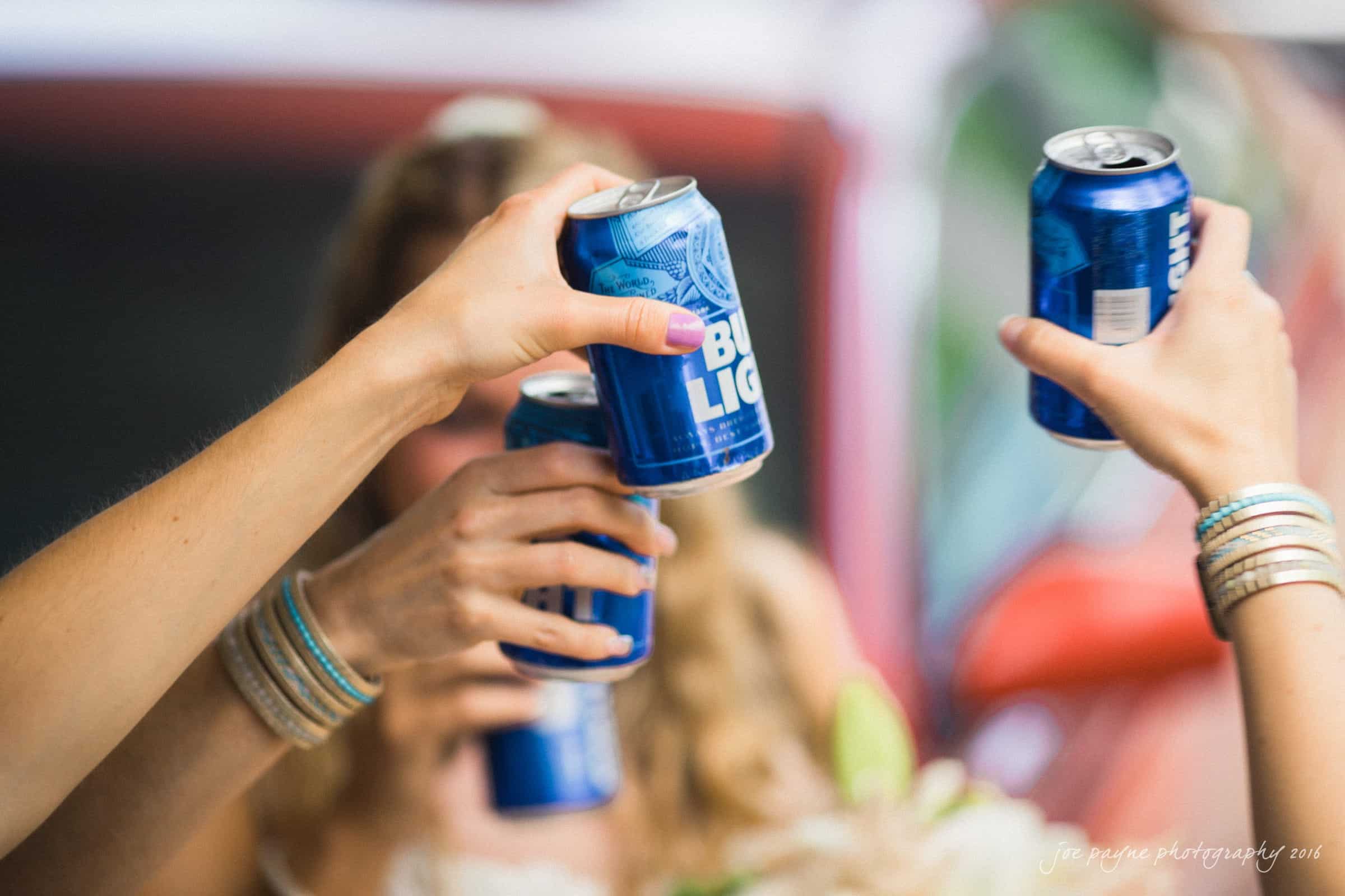 new bern wedding photographer - jessica & brinson Four hands holding Bud Light cans raised in a toast. The background is slightly blurred, showing a hint of a red vehicle. The scene suggests a casual, celebratory moment.