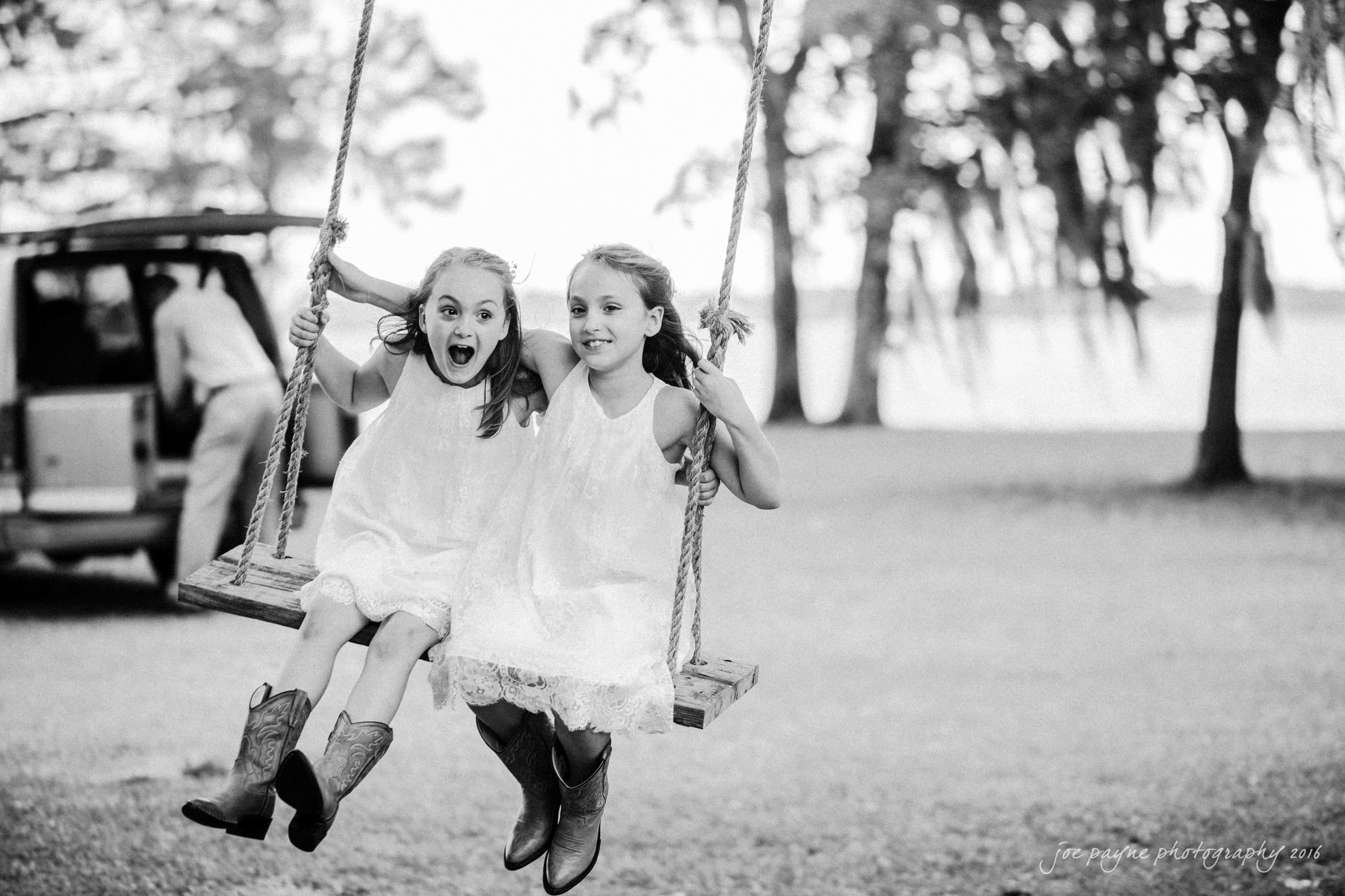 new bern wedding photographer - jessica & brinson Two young girls in white dresses and cowboy boots are sitting on a wooden swing, smiling and making playful expressions. They are outdoors, with trees and a lake visible in the background. A person is near a vehicle in the distance.