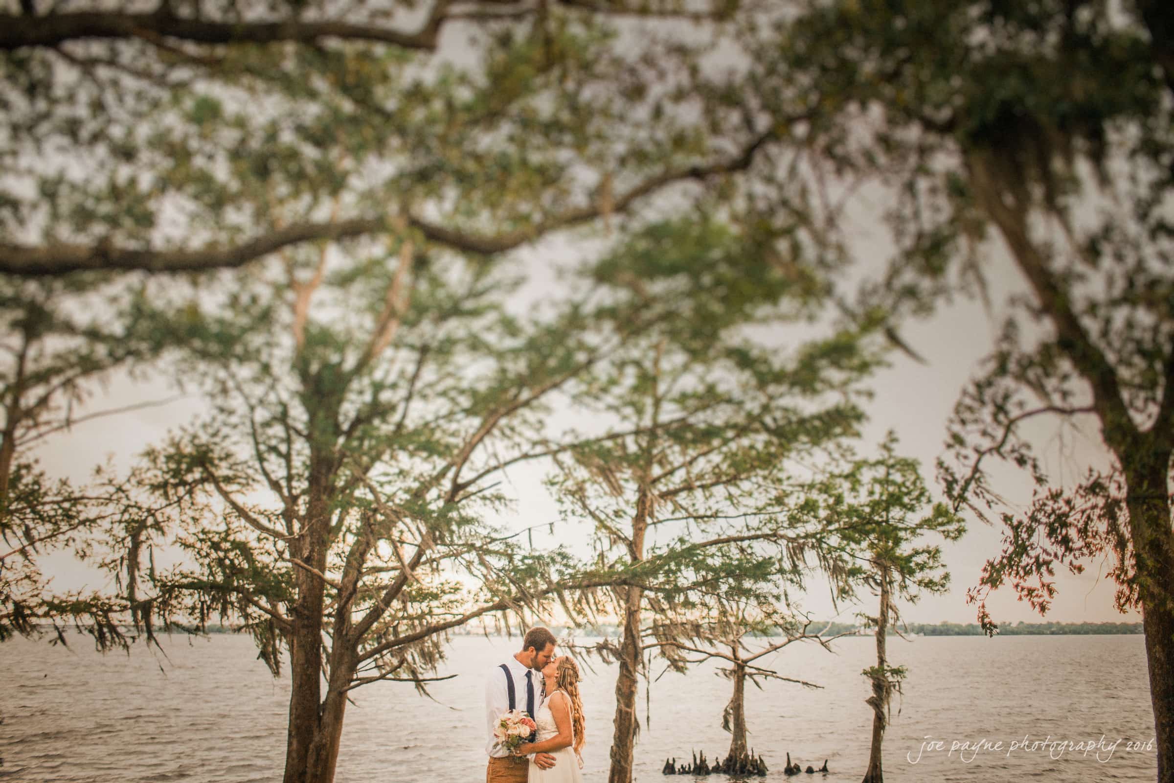 new bern wedding photographer - jessica & brinson A couple dressed in wedding attire shares an intimate moment by a lakeside, surrounded by tall trees. The bride holds a bouquet and they are slightly blurred, adding a dreamy, romantic feel to the scene.