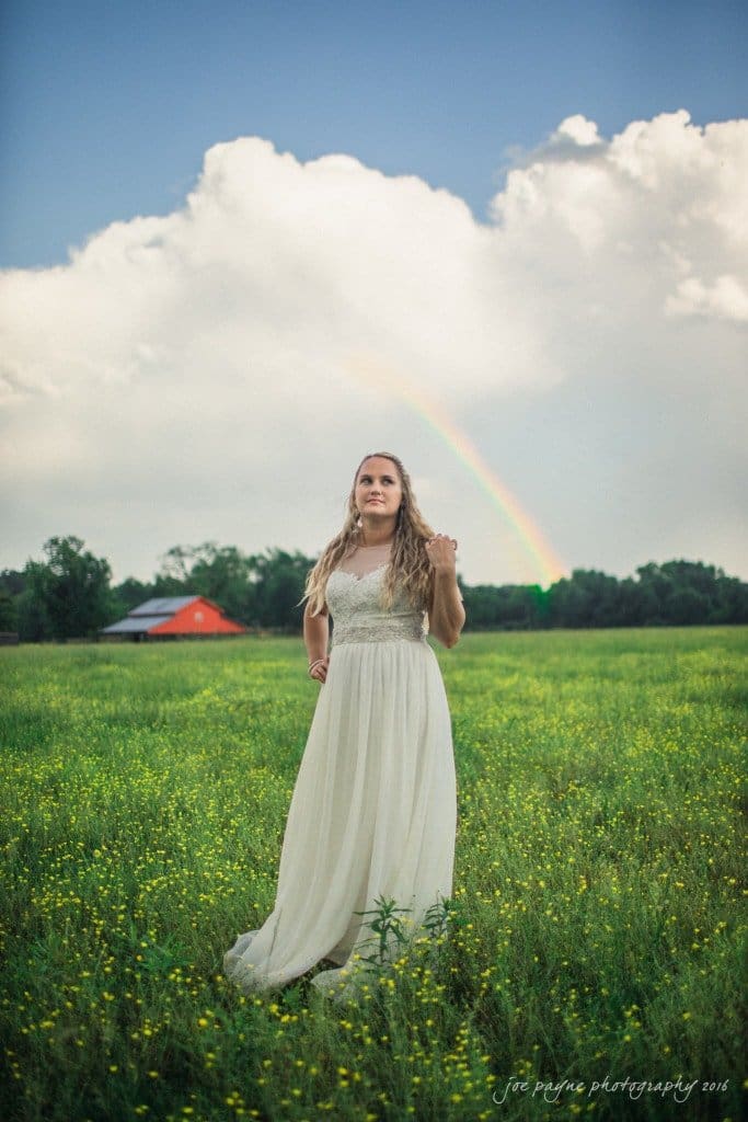 new bern wedding photographer - jessica & brinson A woman in a white dress stands in a field of green grass and yellow flowers. Behind her, a rainbow arches through a partly cloudy sky. A red building is visible in the distant background.