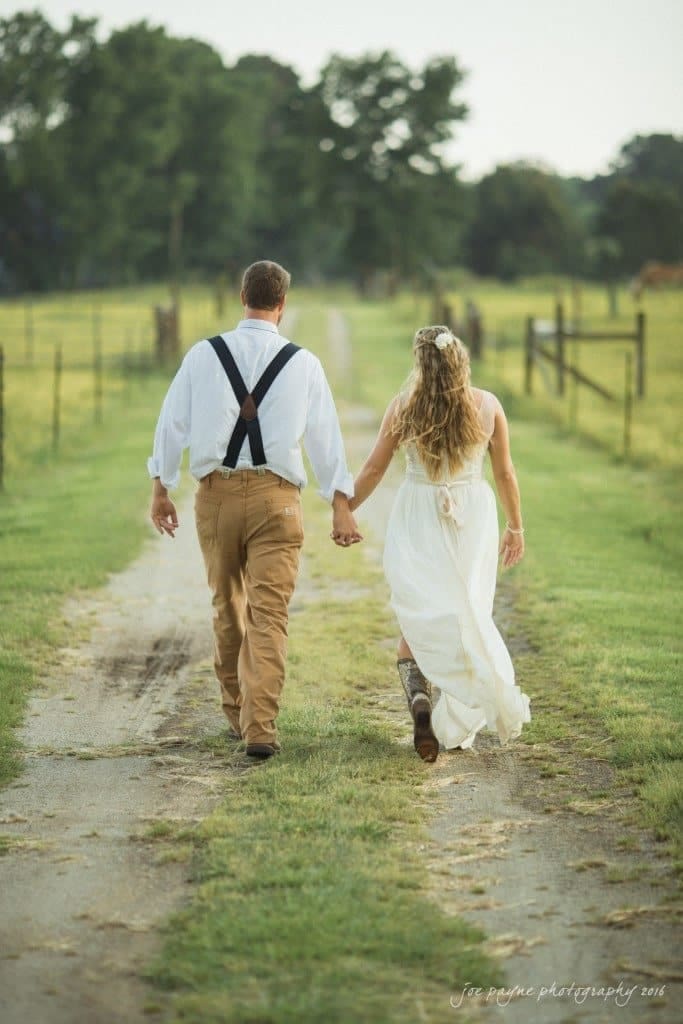 new bern wedding photographer - jessica & brinson A couple walks hand in hand down a country lane. The man wears brown pants, a white shirt, and suspenders, while the woman wears a flowing white dress and cowboy boots. Trees and fencing line the path in the background.