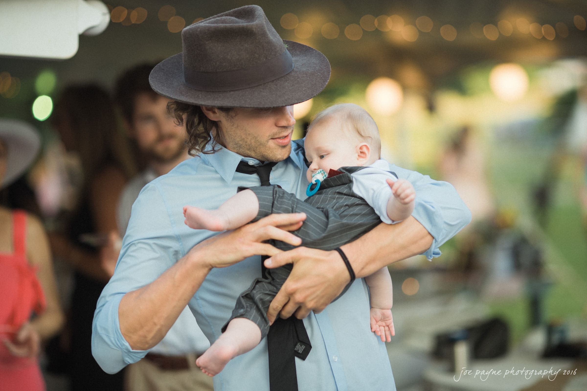 new bern wedding photographer - jessica & brinson A man in a hat and light blue shirt is holding a baby wearing a blue pacifier. The background is slightly blurred with other people and warm lights visible, suggesting a festive outdoor gathering.