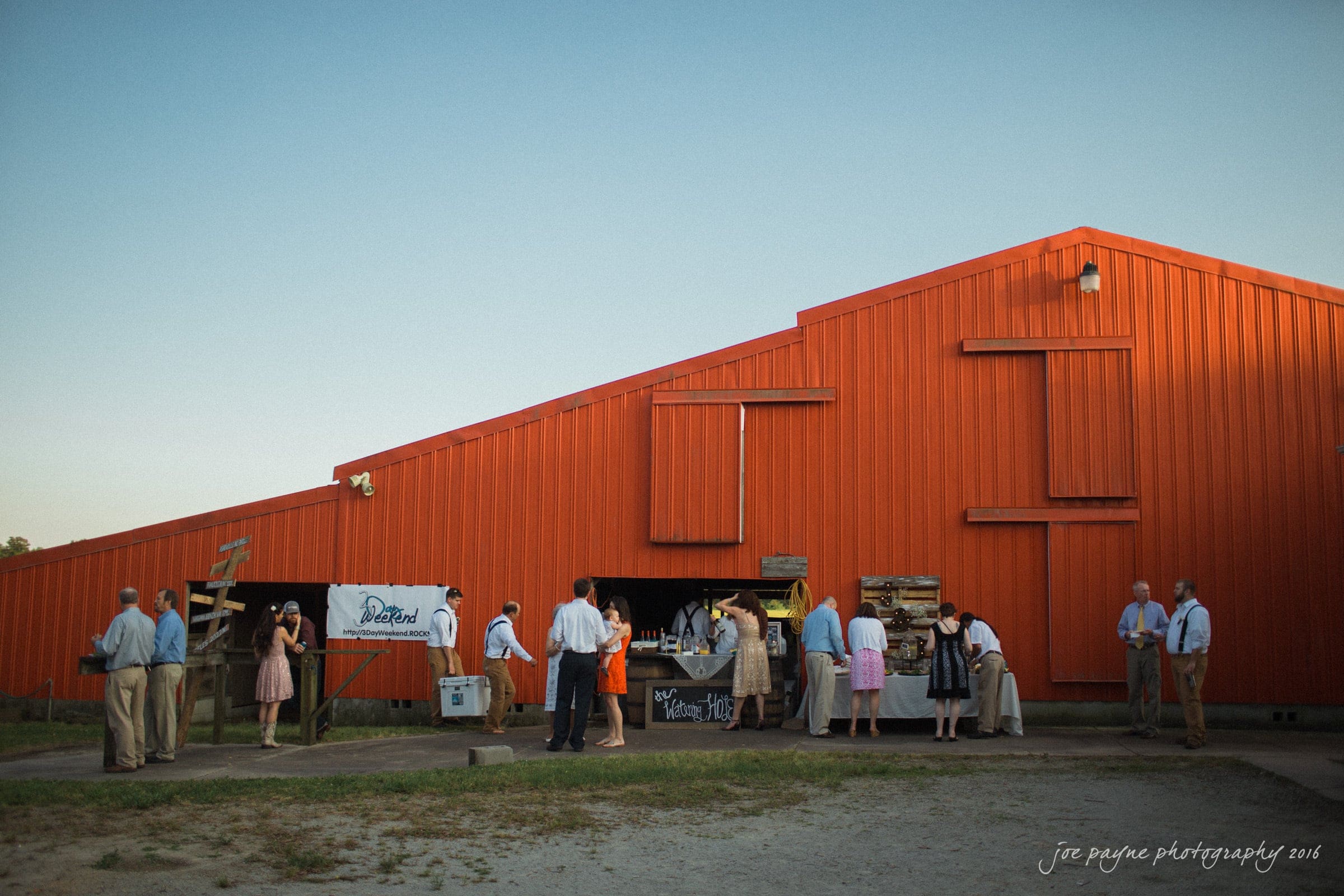 new bern wedding photographer - jessica & brinson A group of people socializing outside a large red barn. Some are gathered near a rustic wooden drink stand, while others chat nearby. The barn contrasts against the clear sky, and the scene has a casual, outdoor gathering atmosphere.