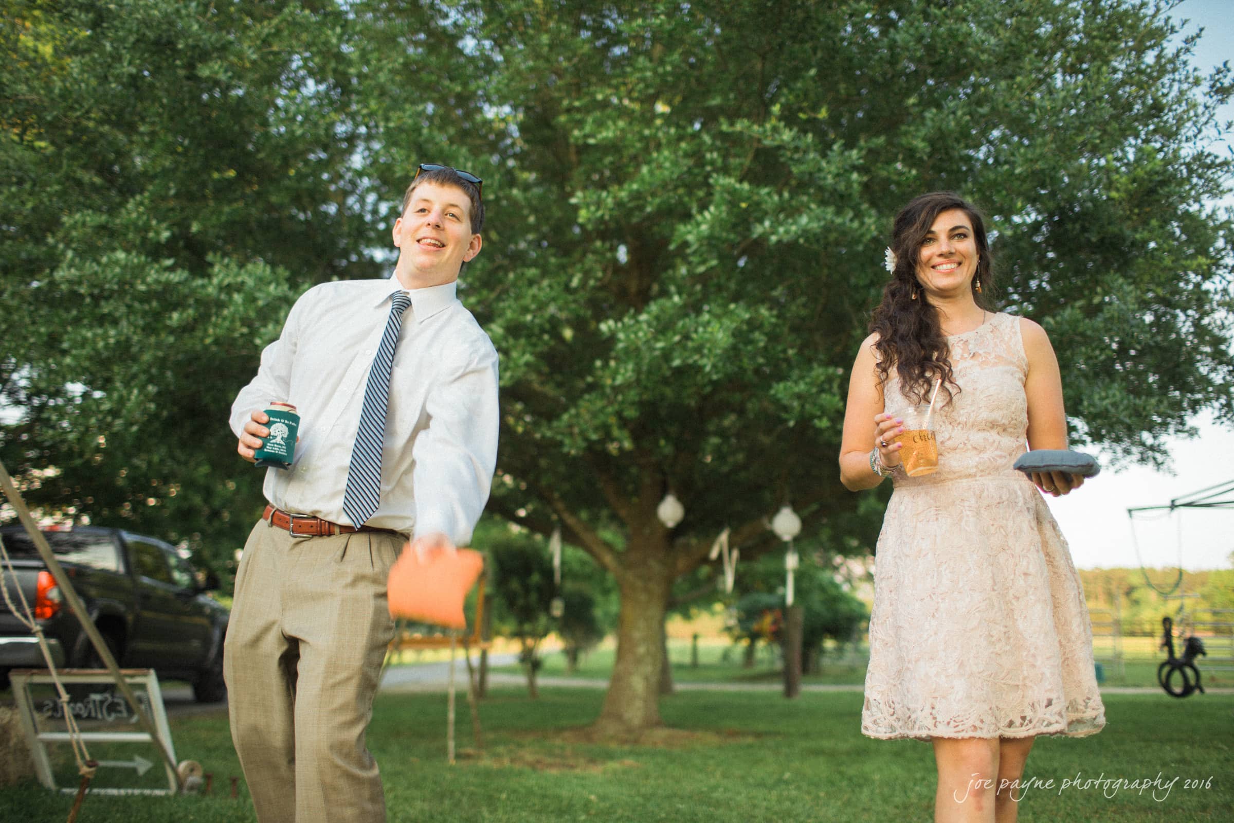 new bern wedding photographer - jessica & brinson A man in a tie and pants tosses a bean bag while holding a drink, and a woman in a lace dress smiles holding a drink and plate. They are outdoors on a lawn with a large tree and blurred background elements.
