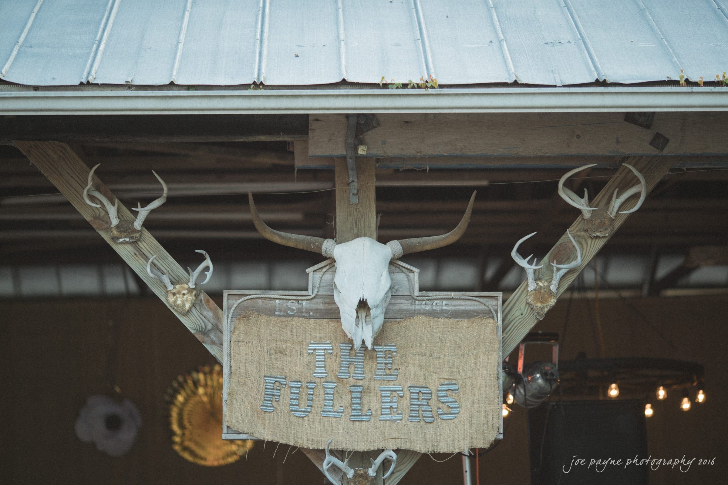 new bern wedding photographer - jessica & brinson A rustic wooden sign with The Fullers hangs under a metal roof. Above it, a cow skull is flanked by several pairs of antlers. The background shows dim lighting and part of a sunflower decoration.