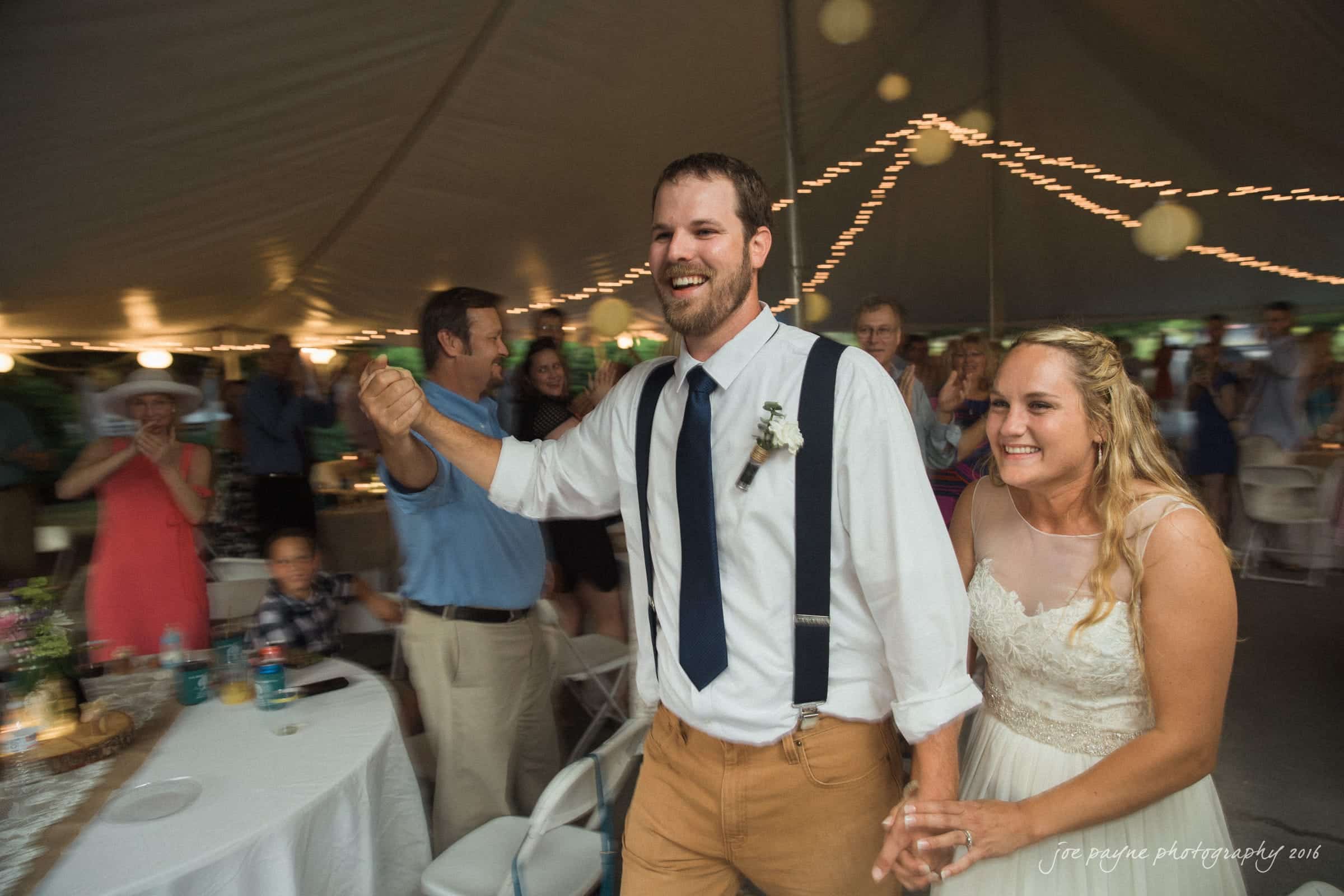 new bern wedding photographer - jessica & brinson A bride and groom, smiling and holding hands, walk into a tented wedding reception. They are followed by cheering guests, with tables and string lights visible in the background. The groom wears suspenders, and the bride is in a white dress.