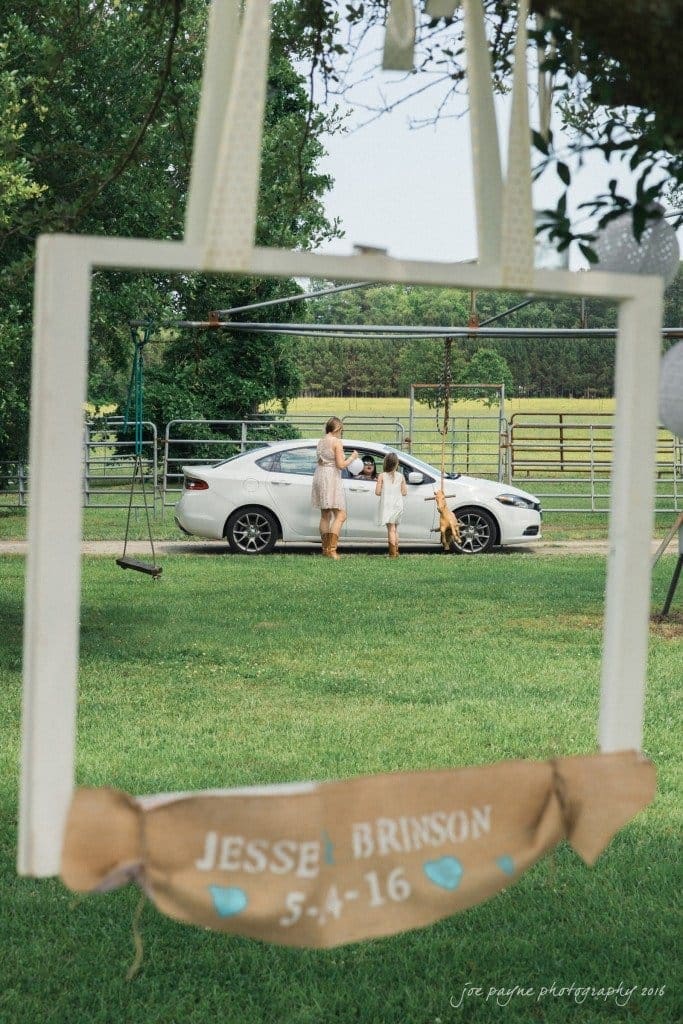 new bern wedding photographer - jessica & brinson A pastoral scene features a white car parked on a lush green lawn. Three people stand near the car, two adults and a child, holding hands. In the foreground, a decorative frame hangs with Jesse Brinson 5-4-16 and small blue hearts.