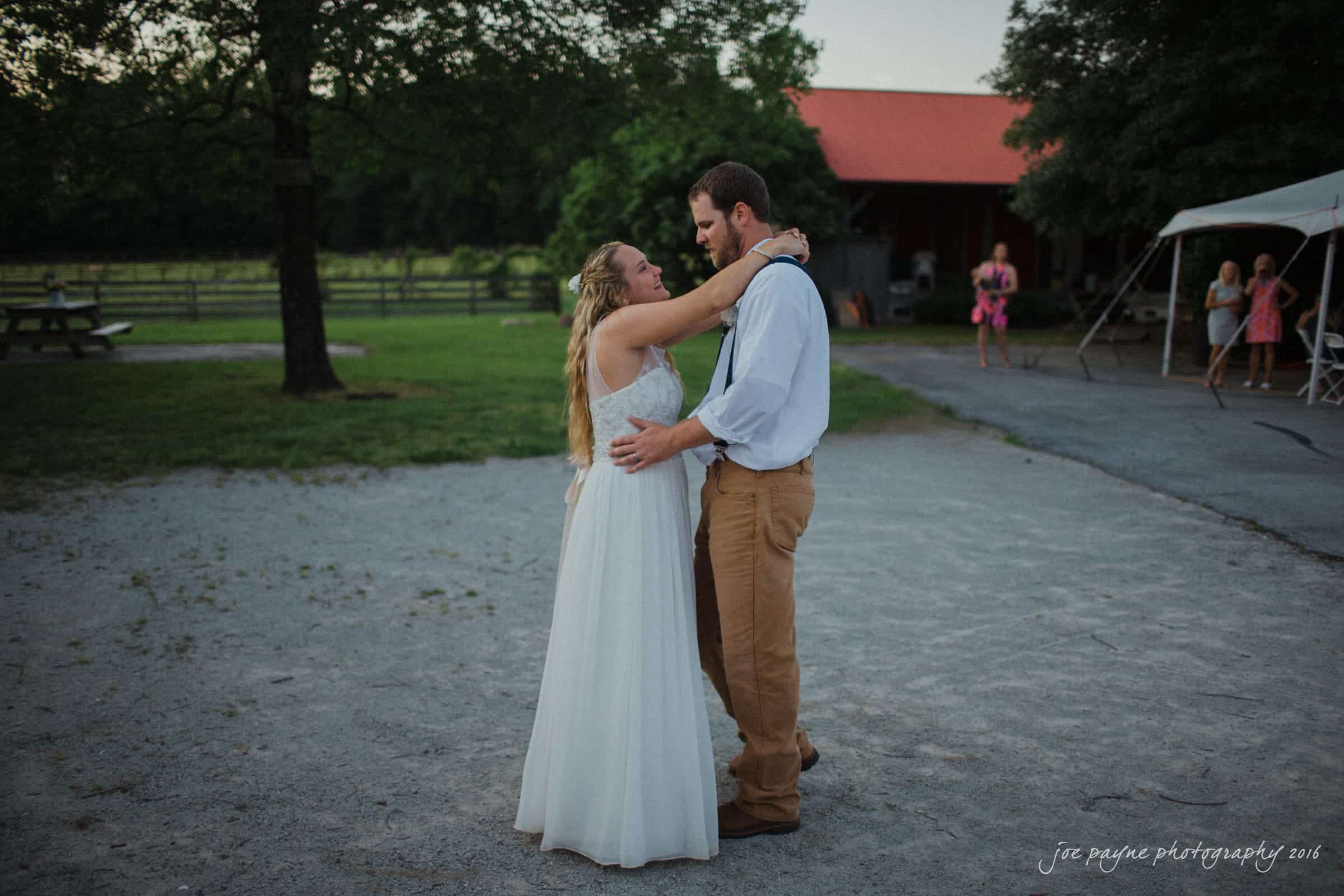 new bern wedding photographer - jessica & brinson A bride in a white dress and a groom in a white shirt and brown pants share a dance outdoors on a gravel path. A red-roofed building and trees are in the background, with a few guests watching from under a canopy.