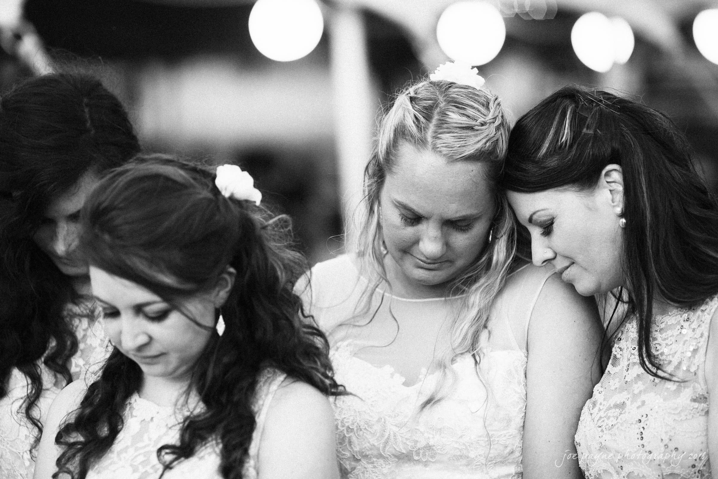 new bern wedding photographer - jessica & brinson A black and white photo of four women standing close together, appearing emotional. They are wearing similar dresses adorned with lace. Two women have their heads bowed, and one rests her head on anothers shoulder.