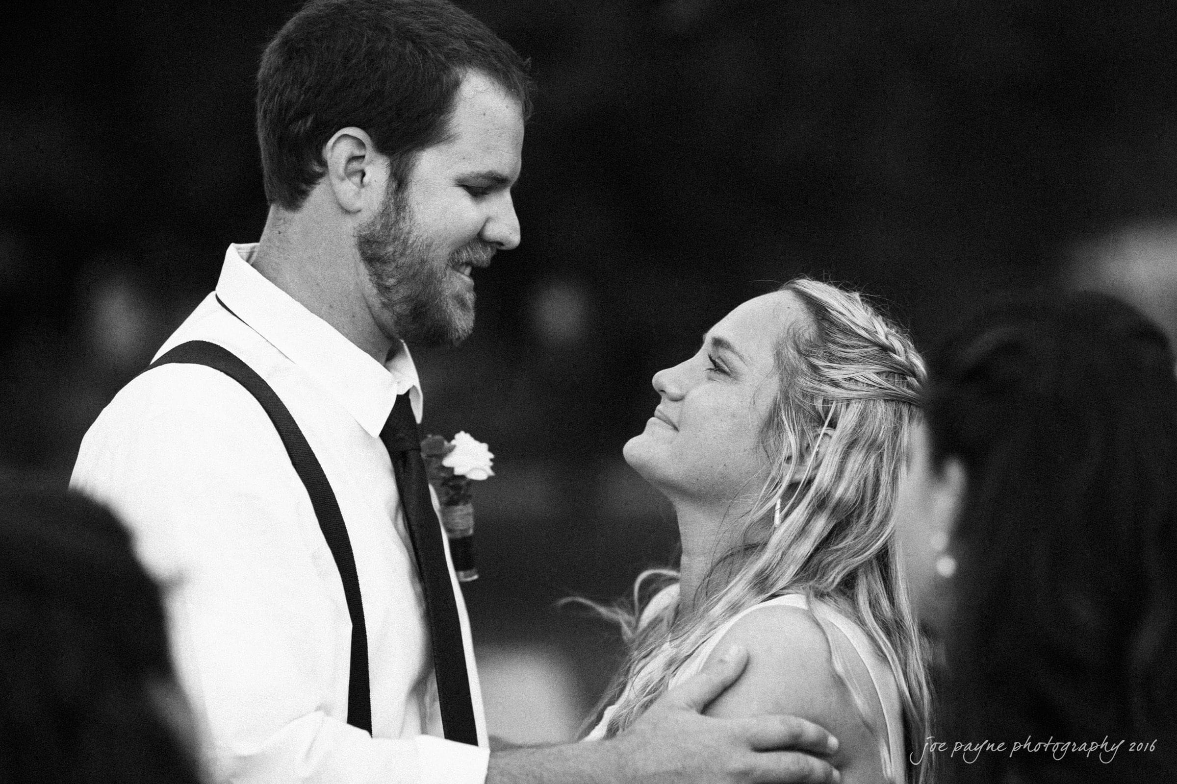 new bern wedding photographer - jessica & brinson A groom and bride share a tender moment, gazing into each others eyes on their wedding day. The groom wears a white shirt and suspenders; the bride has long blonde hair. They are surrounded by guests, and the atmosphere is joyful.