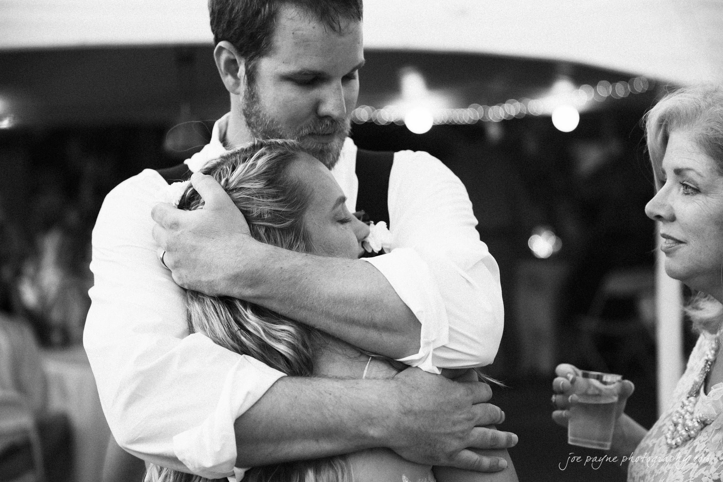 new bern wedding photographer - jessica & brinson A man embracing a woman in a tender hug at an event, captured in black and white. Another woman stands nearby, holding a drink and watching with a gentle expression. The background shows blurred lights and people.