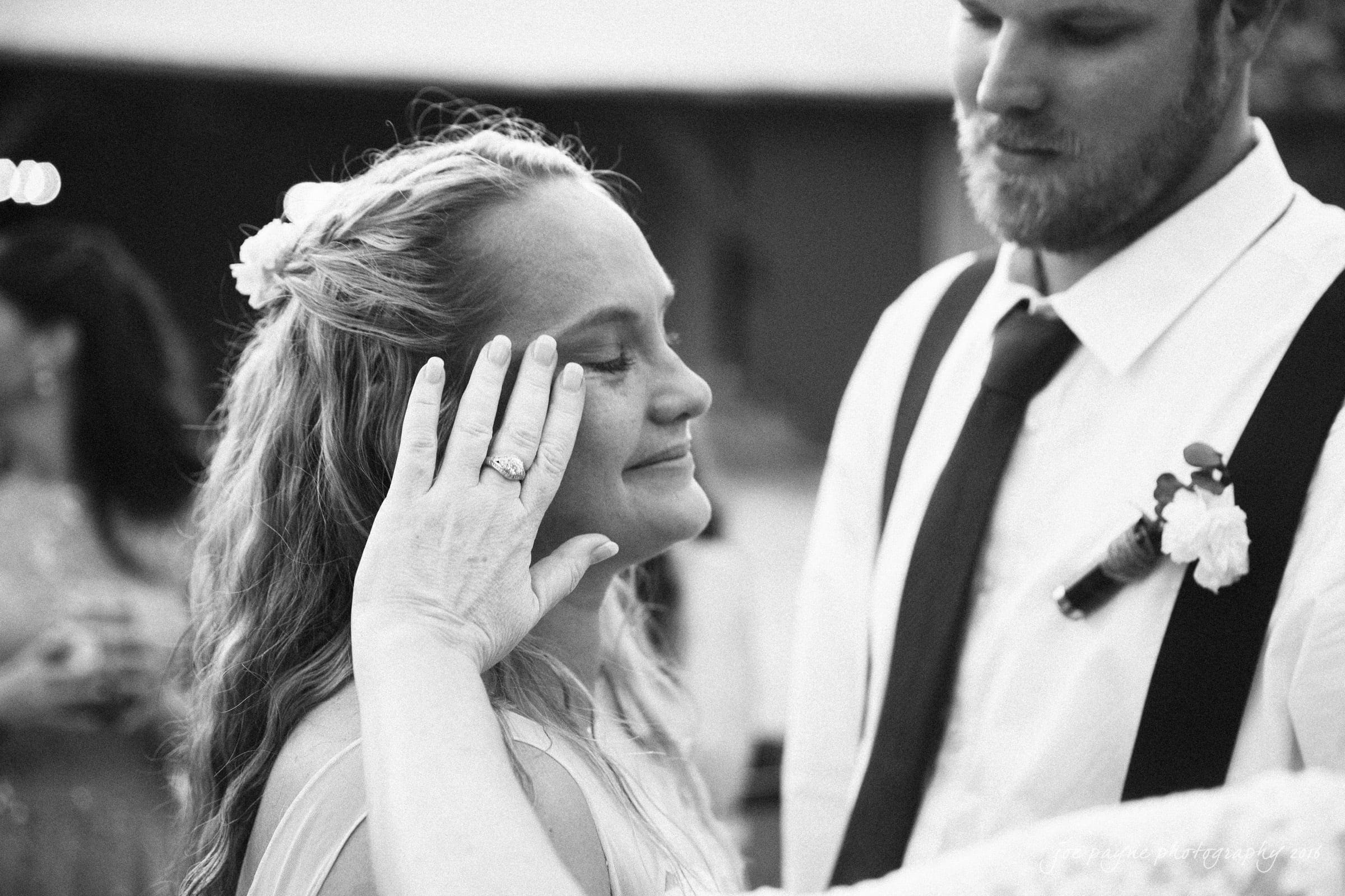 new bern wedding photographer - jessica & brinson A black-and-white photo of a woman with eyes closed, smiling contentedly as a hand touches her face. A man in suspenders stands nearby, gazing at her with a soft expression. Others are blurred in the background.