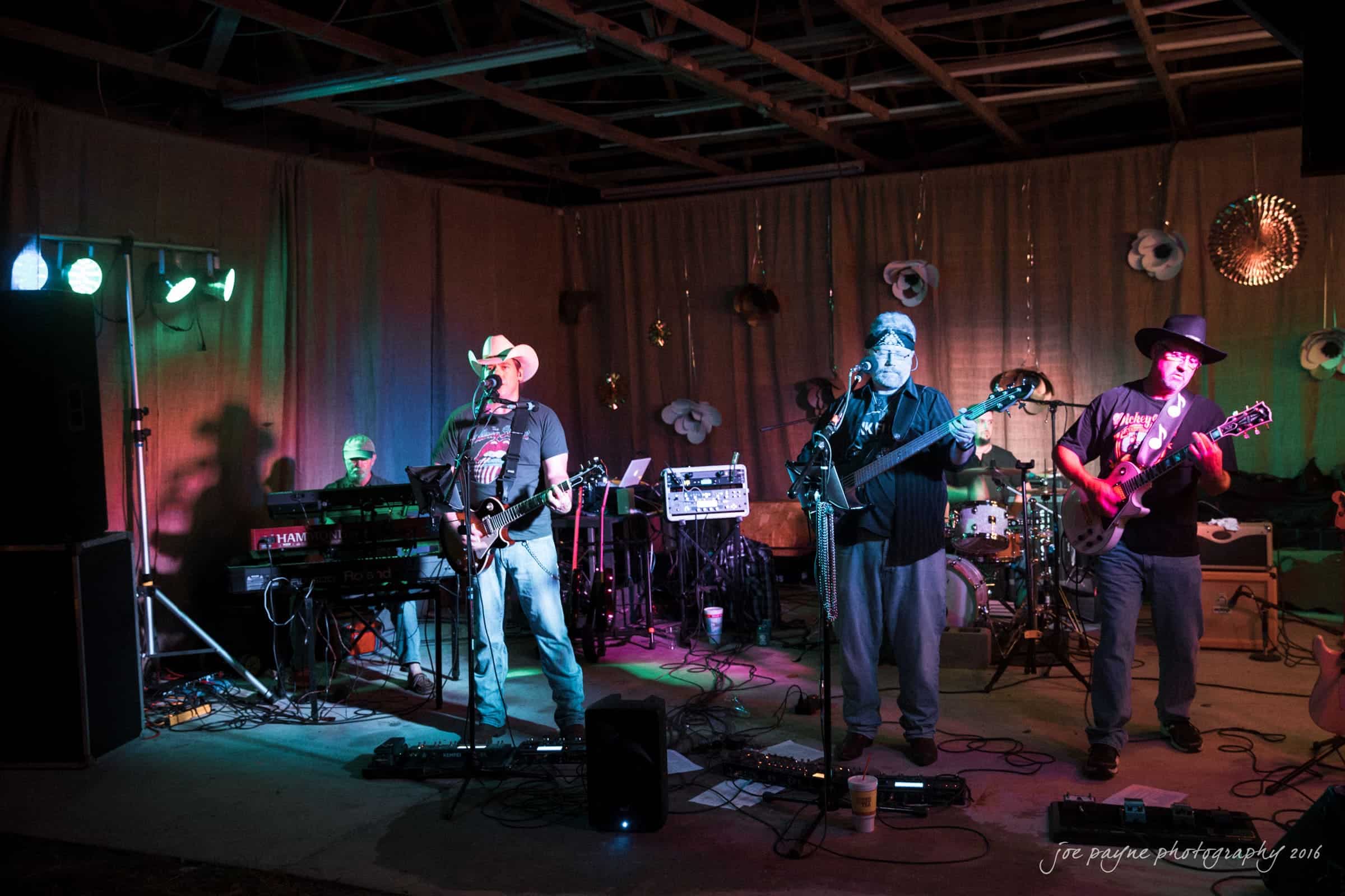 new bern wedding photographer - jessica & brinson A live band performing on a small stage. Three musicians are playing guitar and keyboard under colorful stage lights. The setting has a rustic feel with wooden beams and hanging decorations. The atmosphere is casual and intimate.