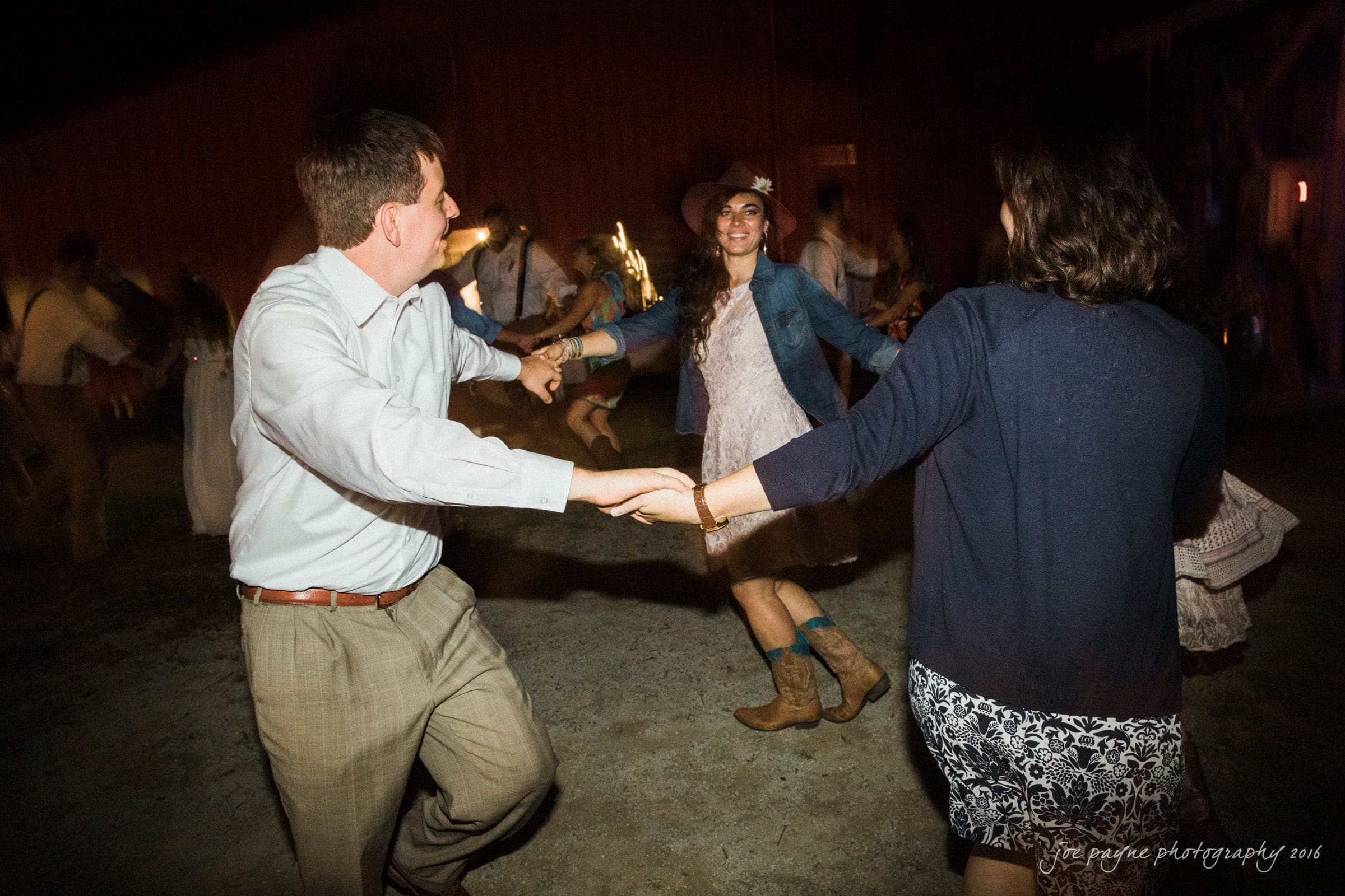 new bern wedding photographer - jessica & brinson A group of people hold hands and dance in a circle at an outdoor event. The woman in the center wears a hat and cowboy boots, smiling brightly. The scene is dimly lit, creating a lively and rustic atmosphere.