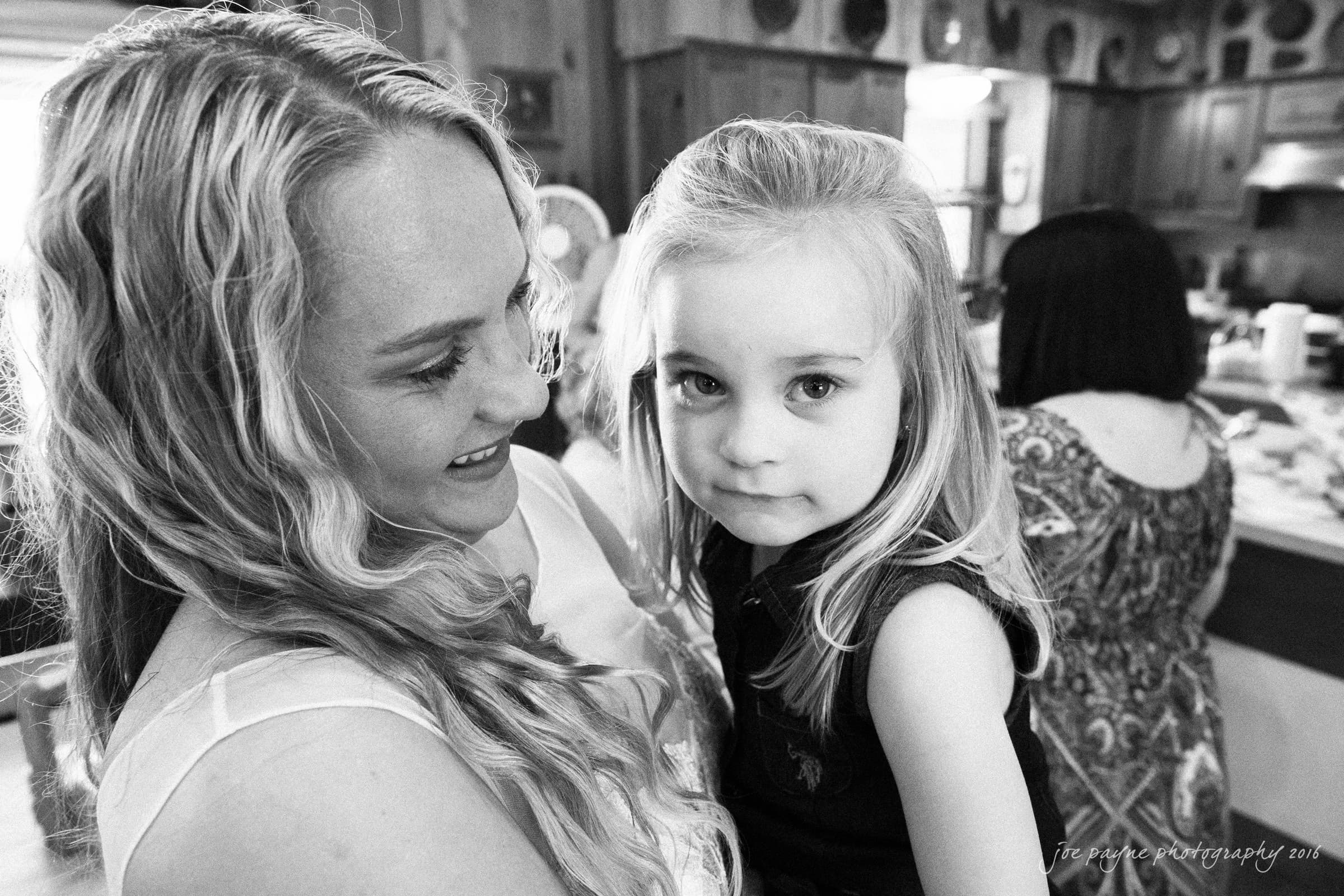 new bern wedding photographer - jessica & brinson A black and white image of a woman with long hair holding a young girl. The woman is smiling at the girl, who looks at the camera with a neutral expression. Another person is in the background in a kitchen setting.