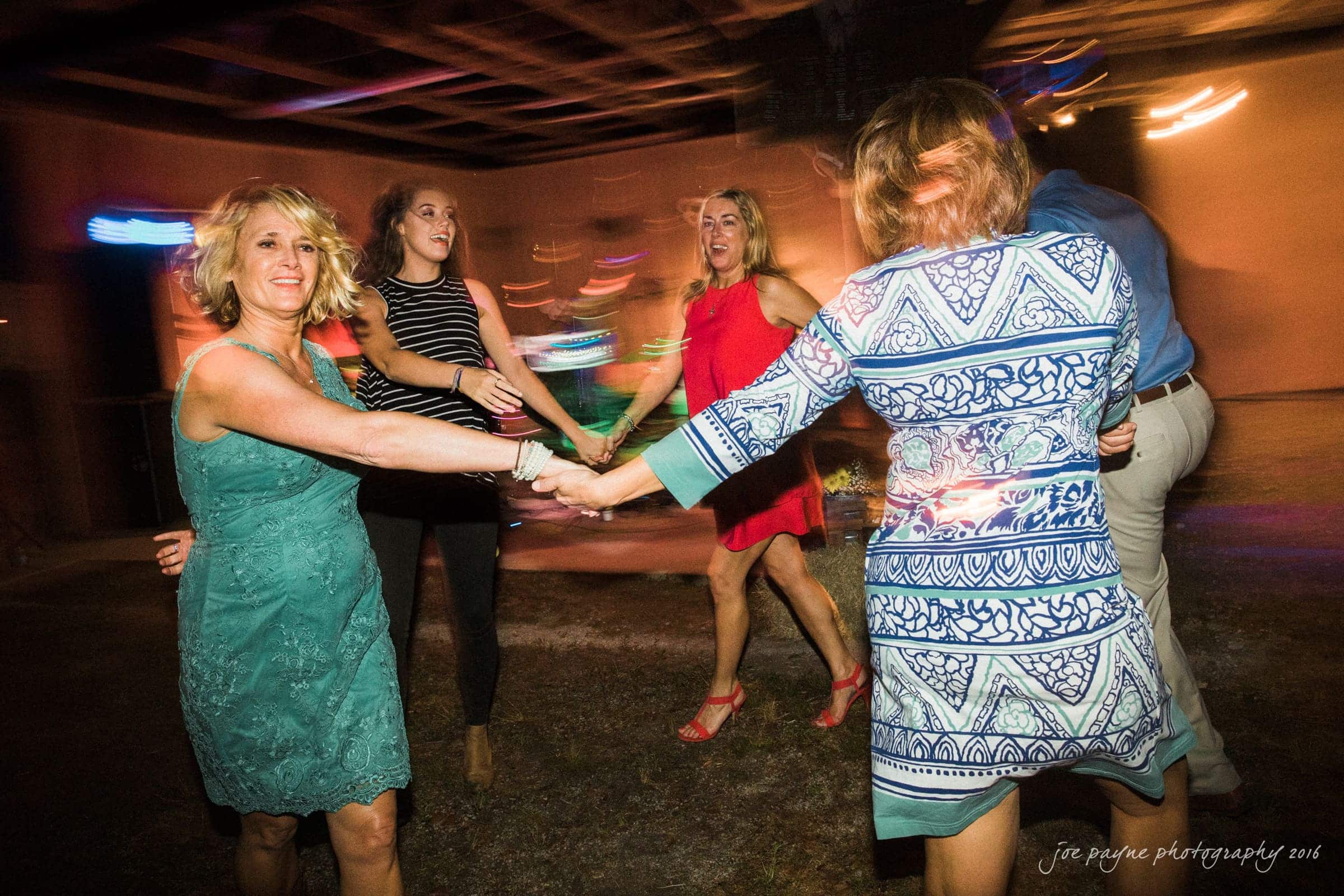 new bern wedding photographer - jessica & brinson A group of people in colorful dresses and outfits form a circle and dance energetically in a dimly lit room with blurred lights. The atmosphere is lively and joyful.