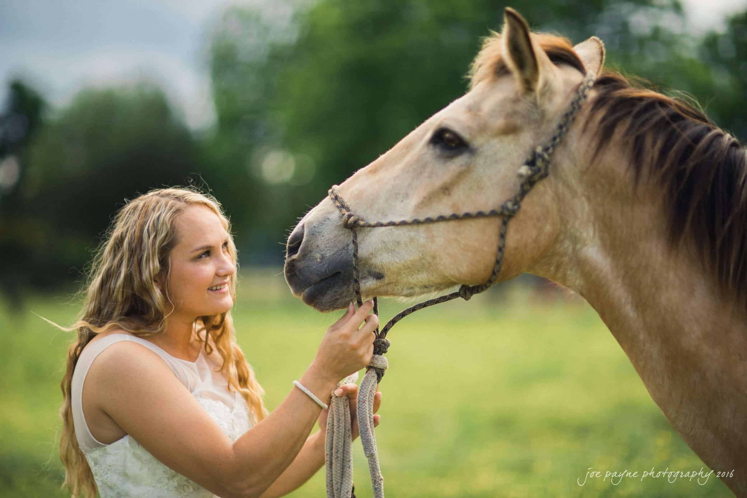 new bern wedding photographer - jessica & brinson A person with long curly hair, wearing a sleeveless top, gently holds a rope around a horses neck. They are outdoors in a lush, grassy area with trees in the background.