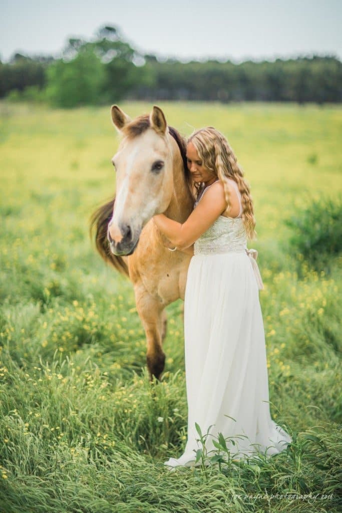 new bern wedding photographer - jessica & brinson A woman in a white dress lovingly embraces a horse in a lush green field with yellow wildflowers. Trees are visible in the background.
