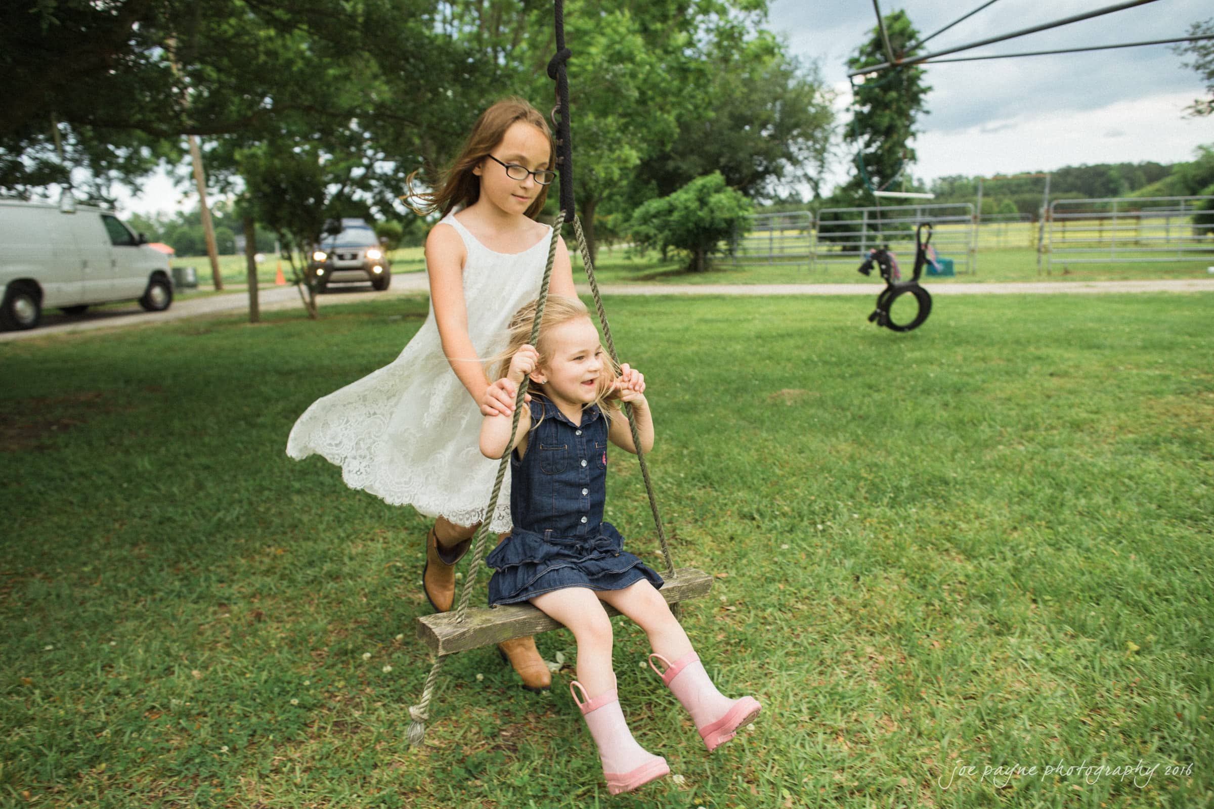 new bern wedding photographer - jessica & brinson Two young girls playing on a wooden swing in a grassy yard. The older girl is wearing a white dress, pushing the younger girl in a denim dress and pink boots. Trees, a tire swing, and vehicles are visible in the background.