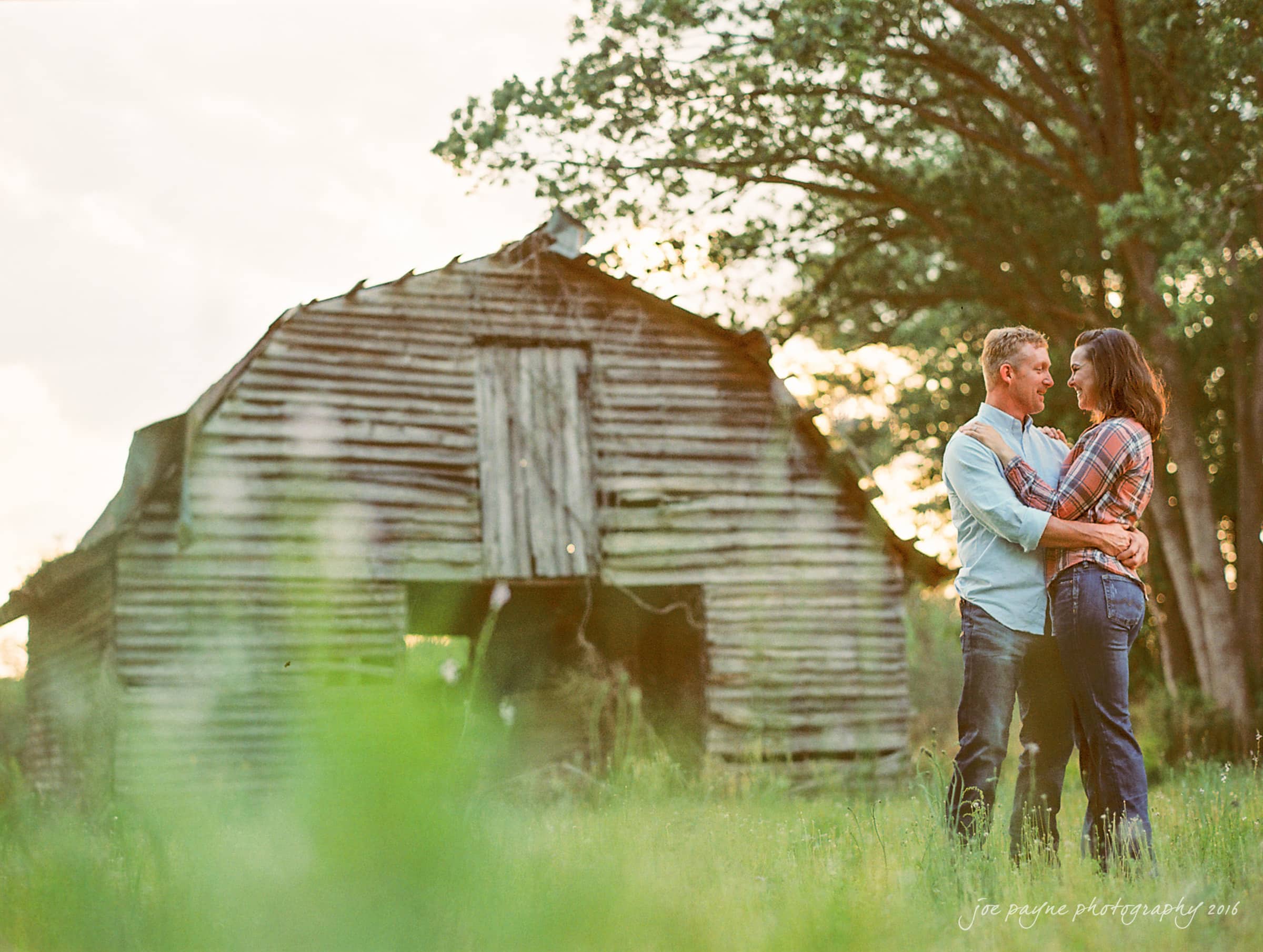 A couple stands closely, embracing and smiling at each other, in front of a rustic wooden barn surrounded by grass and trees, under a soft, cloudy sky.