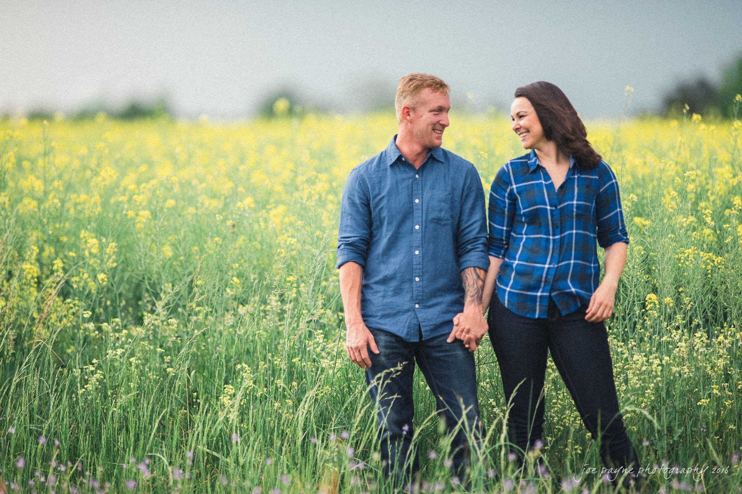 A couple holding hands, walking through a lush field of yellow flowers. The man is wearing a blue shirt, and the woman is in a blue plaid shirt. They are smiling at each other, surrounded by greenery under a cloudy sky.