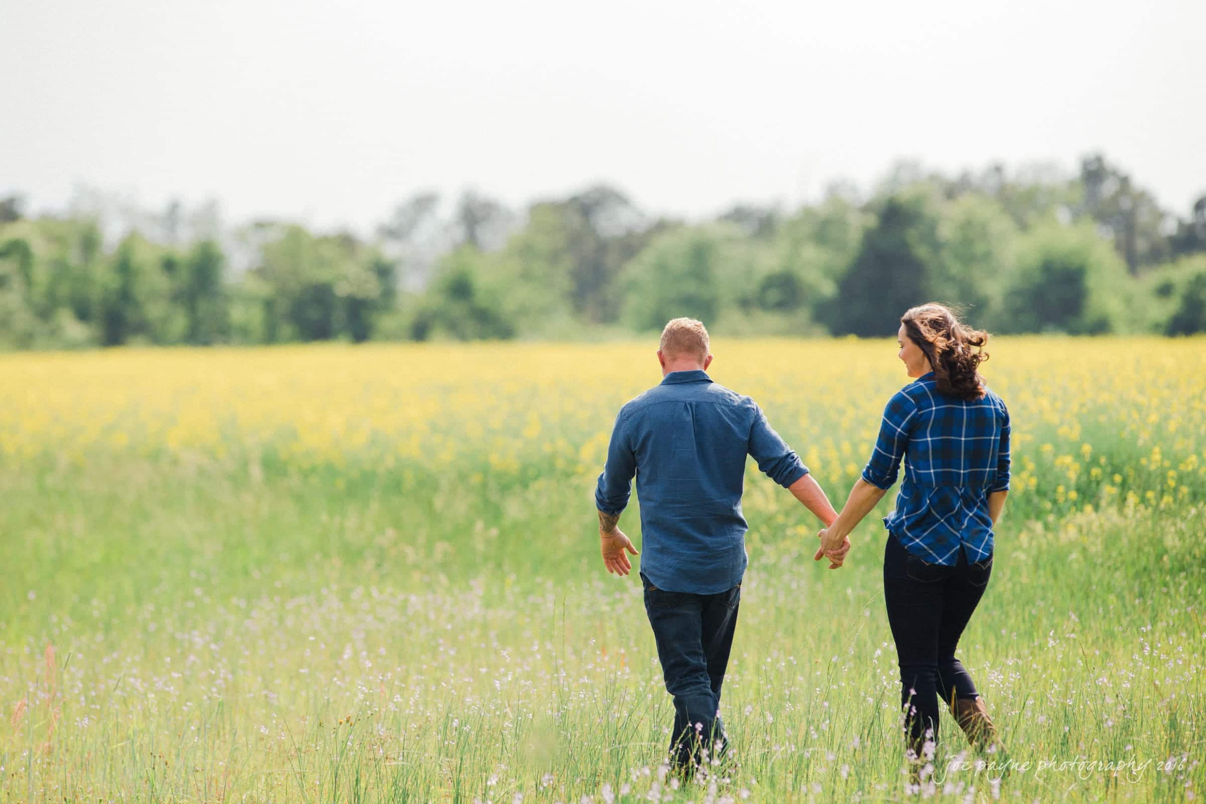 A couple holding hands and walking through a vibrant green field with tall grass and yellow wildflowers, surrounded by distant trees under a clear sky. Both wear blue shirts, adding to the peaceful rural scene.