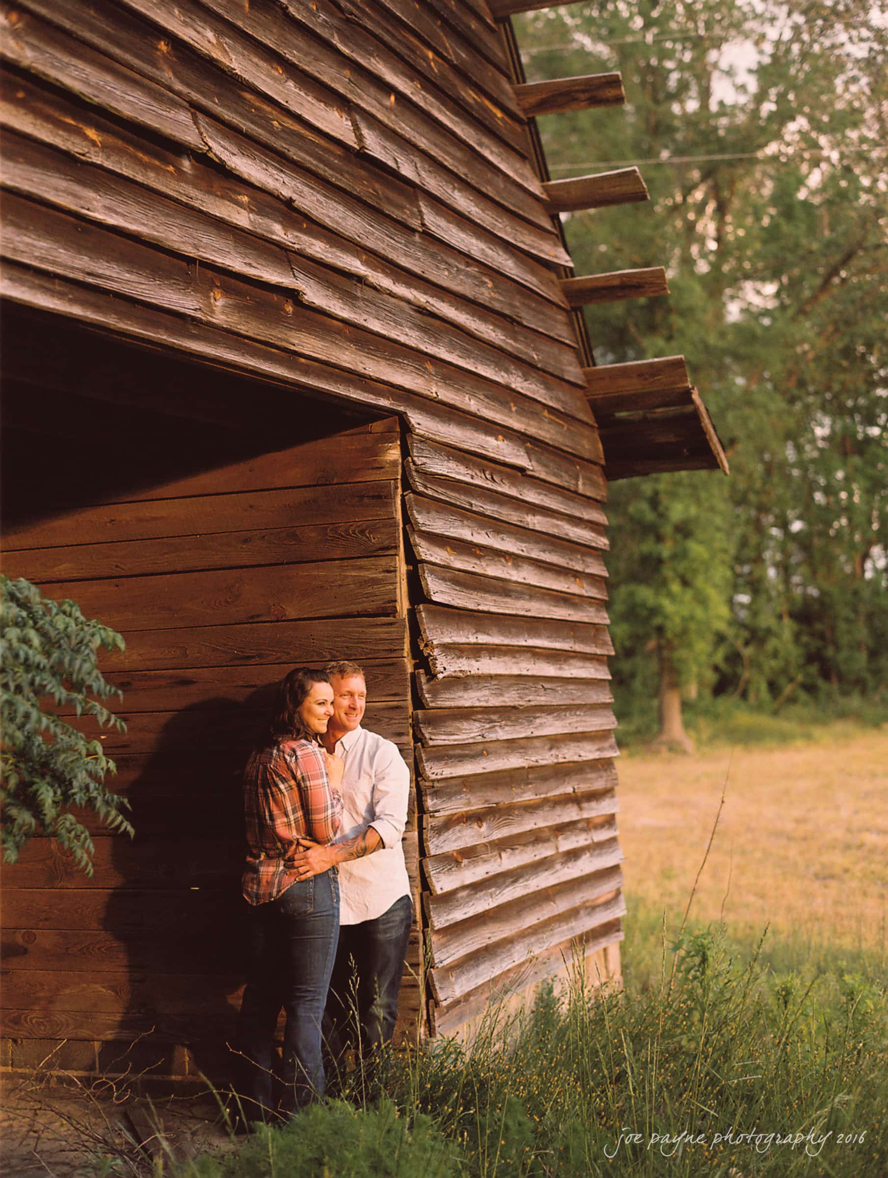 A couple embraces and smiles while standing against the rustic wall of a wooden barn. They are surrounded by tall grass, and trees are visible in the background. The lighting is warm and gives the scene a serene, romantic atmosphere.