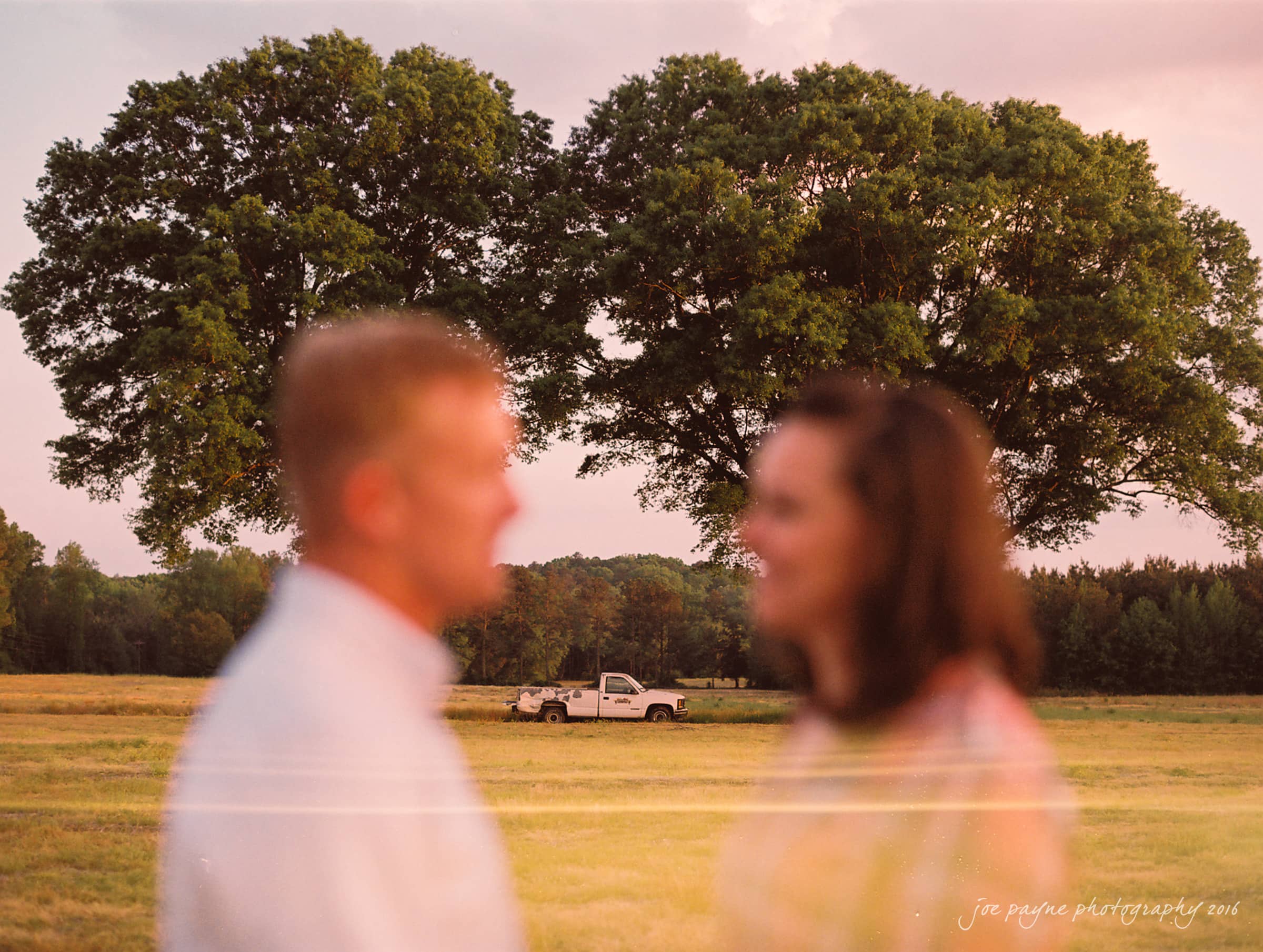 A couple is standing in a grassy field with the focus blurred. In the background, a white pickup truck is parked under two large trees. The scene is lit by soft, warm lighting during sunset.