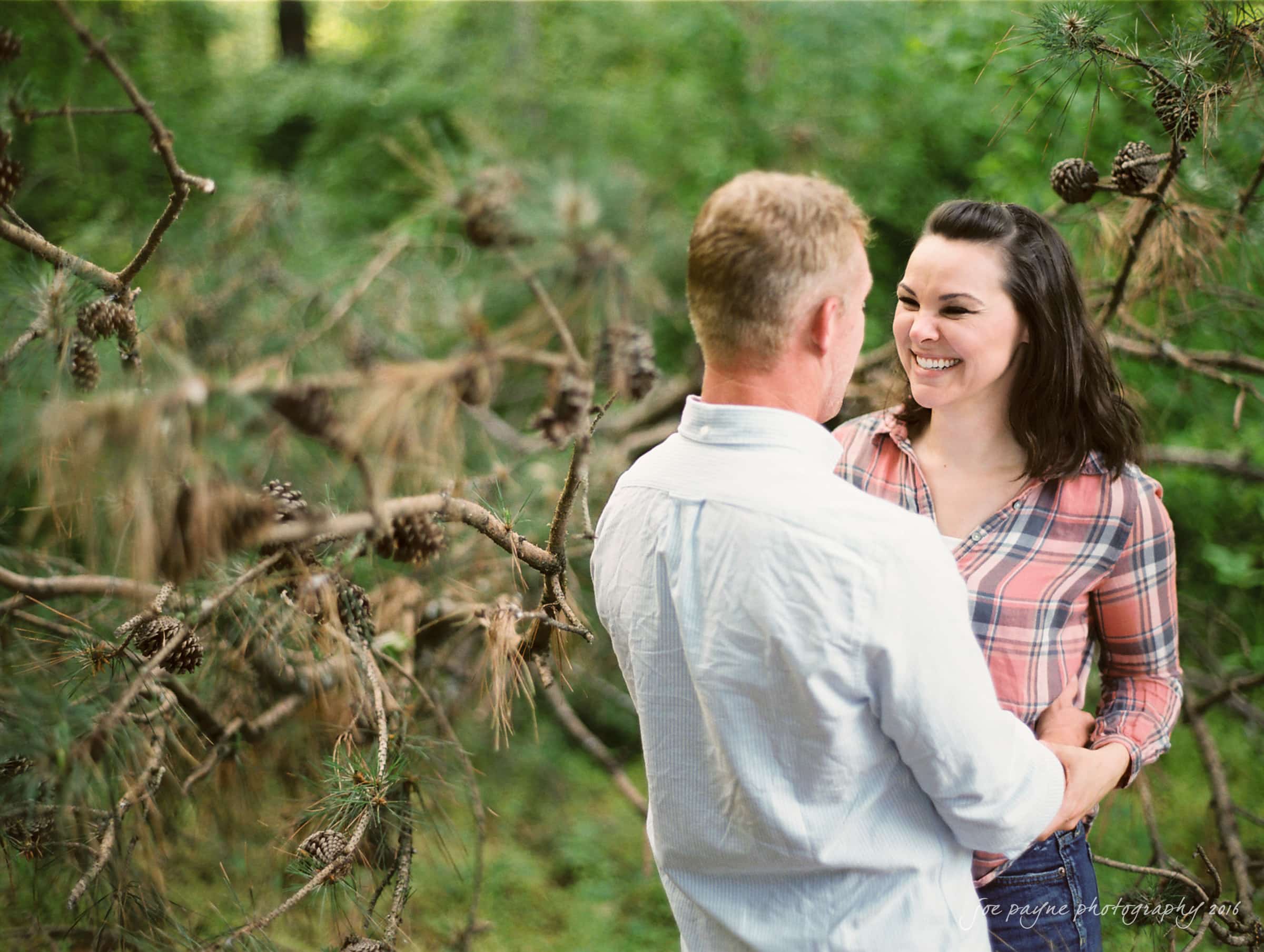 A couple stands close together, smiling at each other, surrounded by a lush forest backdrop. The man faces away from the camera in a white shirt, and the woman wears a plaid shirt, her hair loose. They appear happy and relaxed.