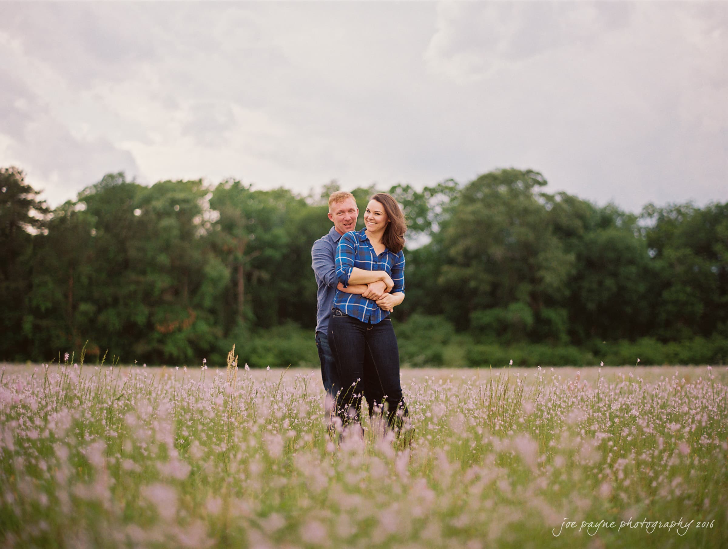A couple embracing and smiling in a field of tall grass and pink flowers, with a background of green trees and a cloudy sky.