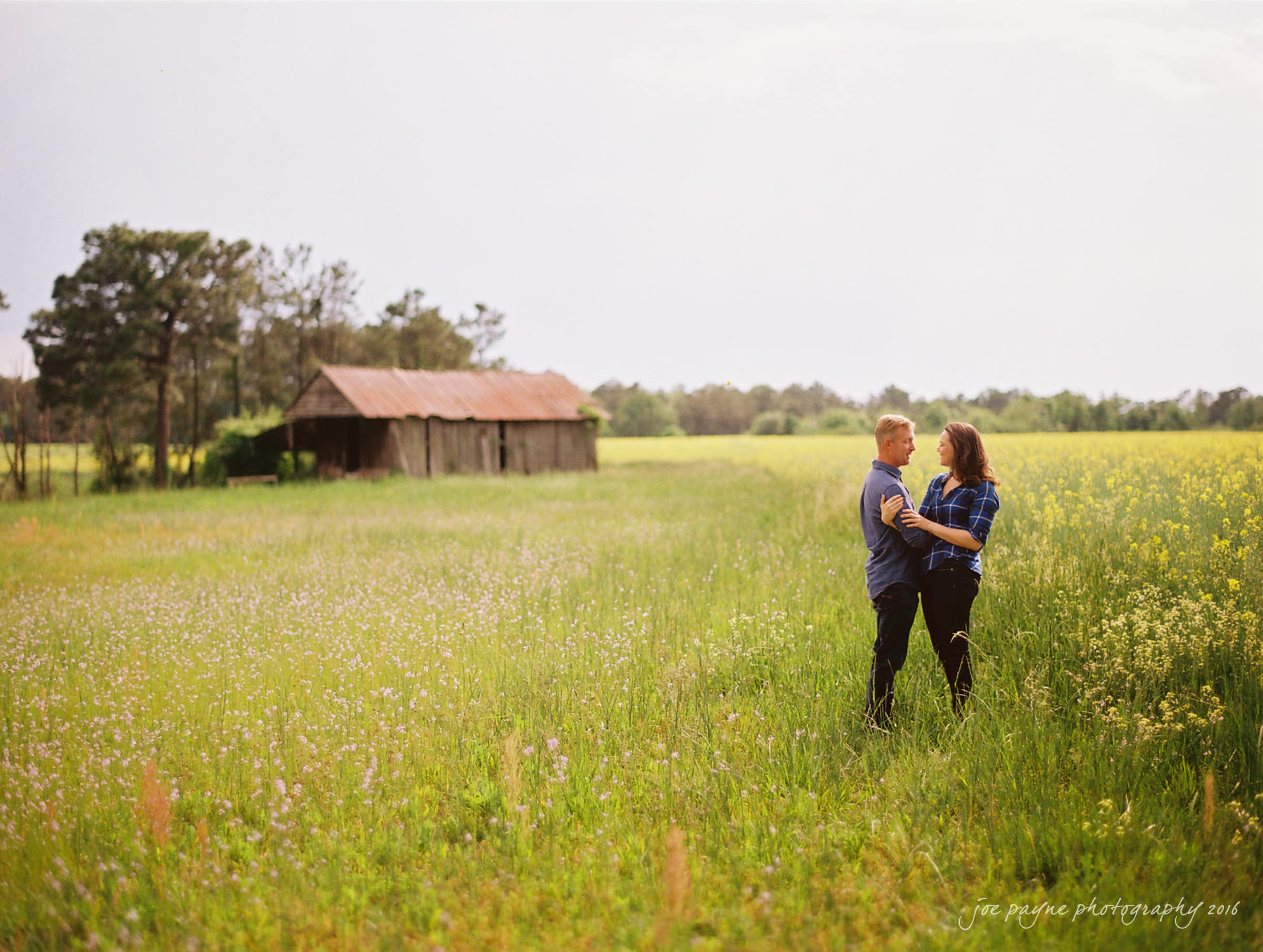 A couple stands in a field of wildflowers, embracing and smiling at each other. A rustic barn and trees are in the background under a light, cloudy sky.