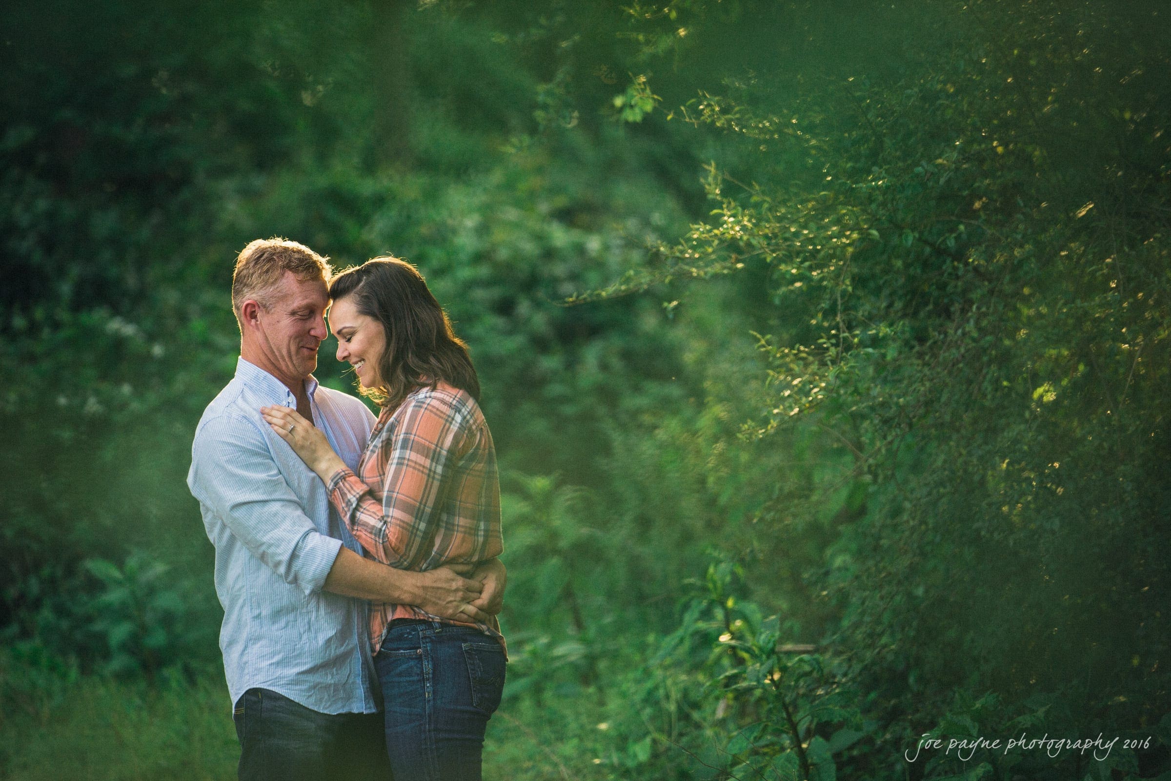 A couple embraces in a lush, green outdoor setting. The man wears a light blue shirt, and the woman wears a plaid shirt. Sunlight filters through the trees, creating a warm, tranquil atmosphere.