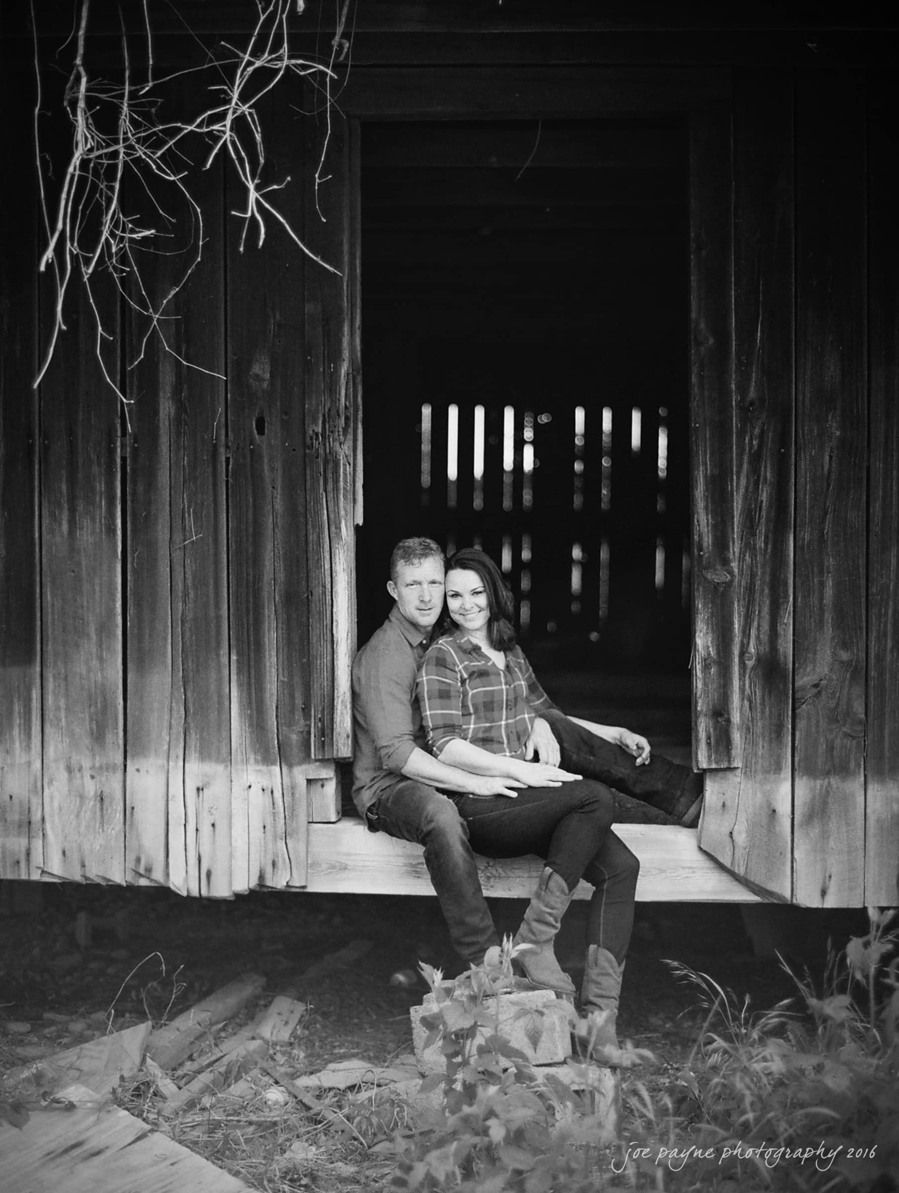 A couple sits in a wooden barn doorway, both smiling. They are wearing casual clothes. The barns interior is dark, creating a contrasting backdrop. Some foliage is visible around the barn.