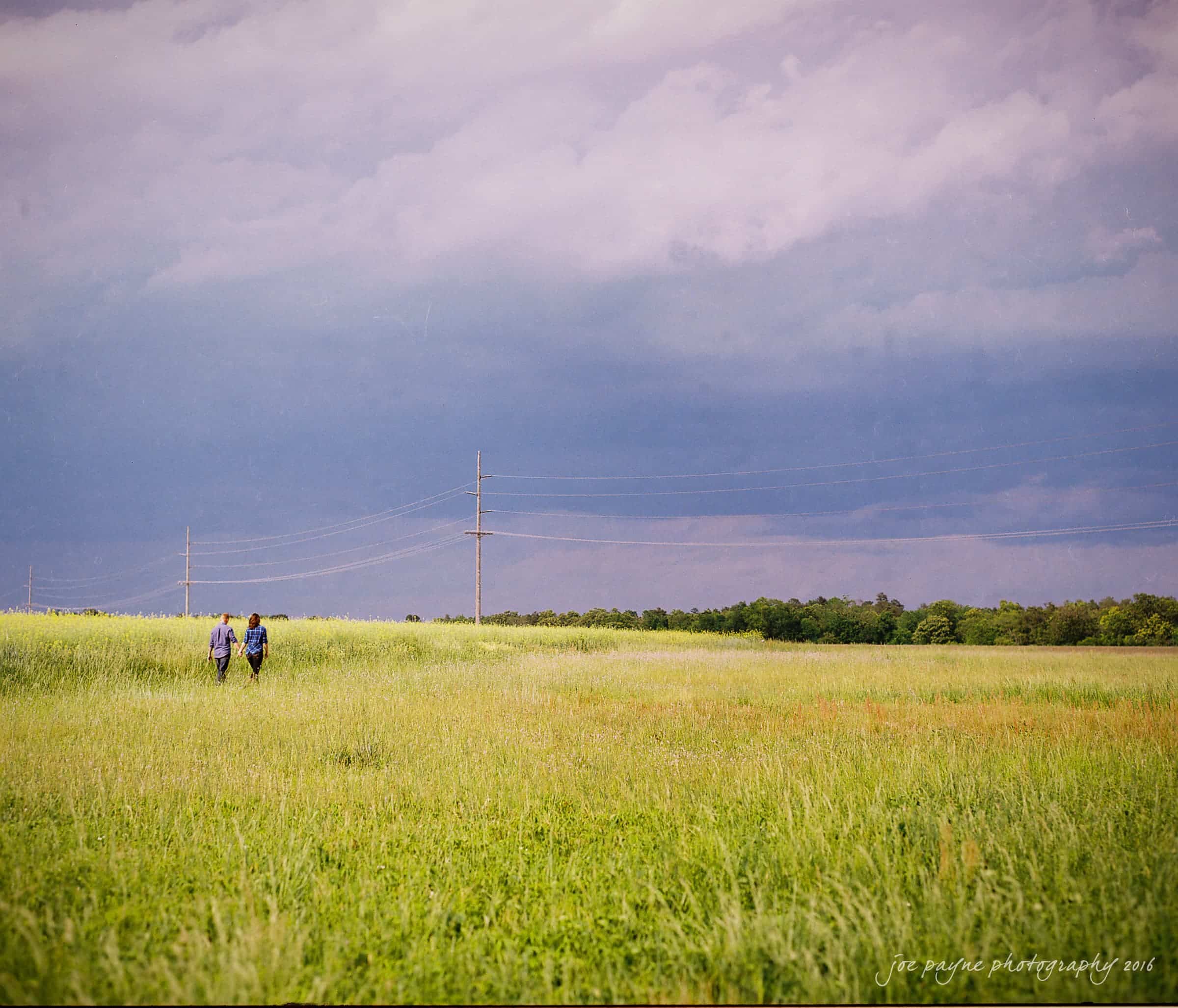 Two people walk through a vast green field under an expansive, moody sky with scattered clouds. Power lines stretch across the horizon, and trees line the distant background, adding depth to the serene landscape.