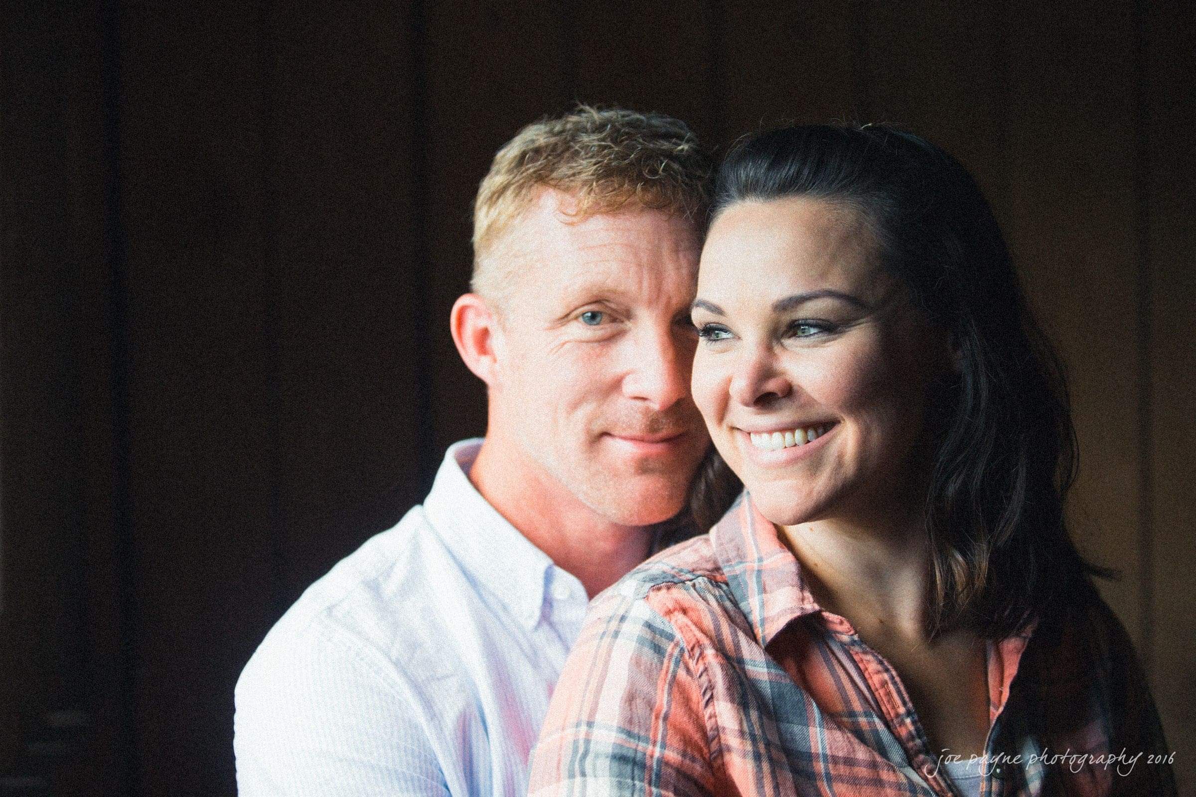 A couple sits closely together, smiling warmly. The man is wearing a white shirt while the woman is dressed in a plaid shirt. Soft, natural light highlights their expressions against a dark background.