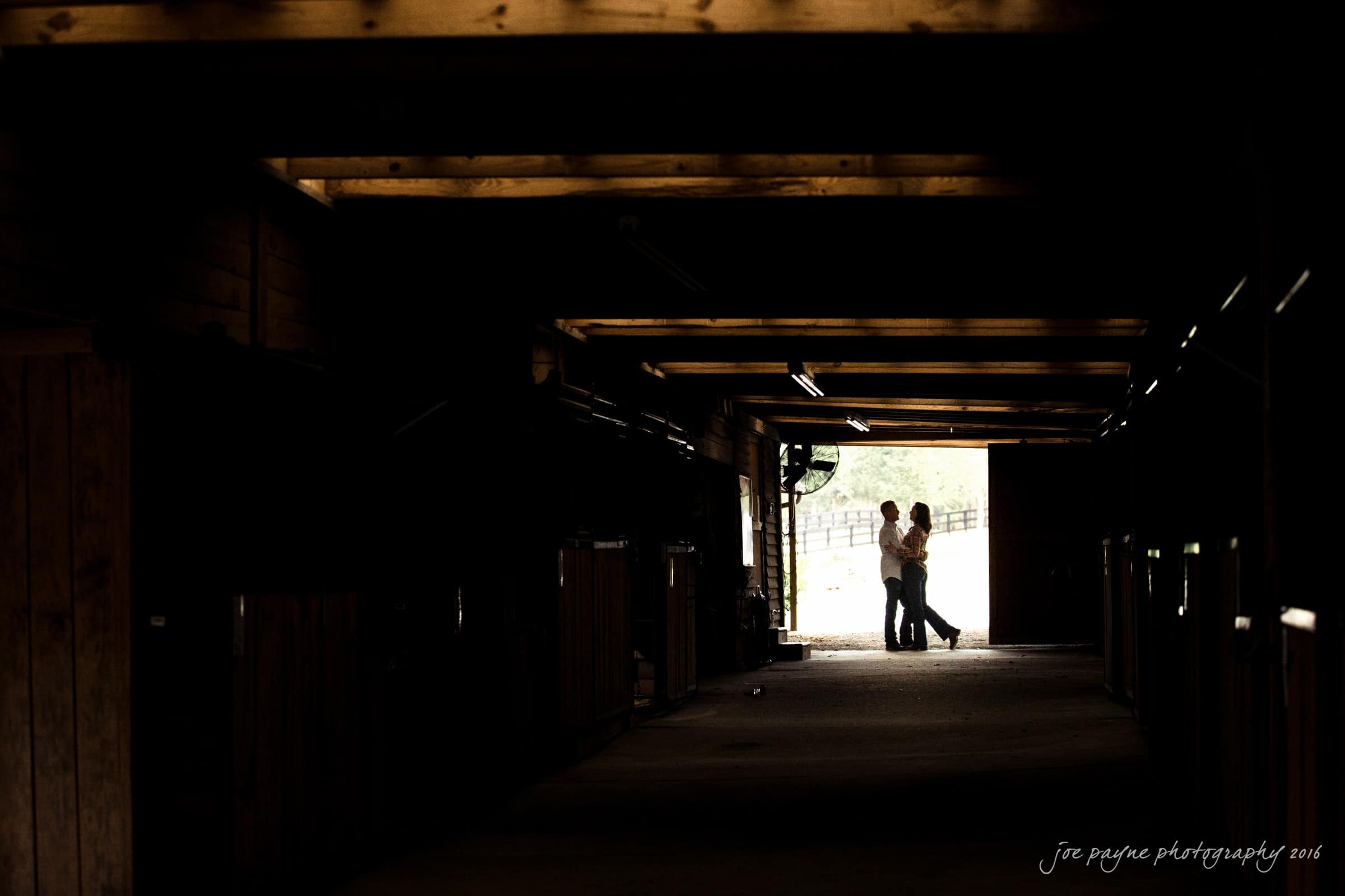 A couple stands in a barn doorway, silhouetted against the light outside. The dimly lit interior features wooden beams and horse stalls. They embrace, surrounded by the rustic charm of the barn setting.