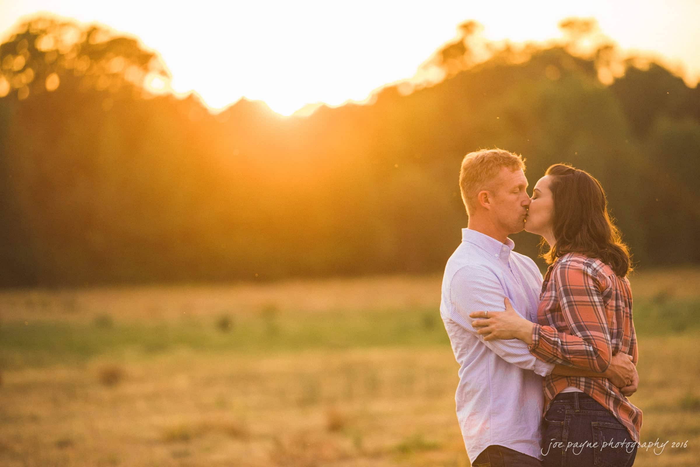 A couple embraces and kisses in a sunlit field during sunset. The man wears a light blue shirt, and the woman is in a plaid shirt. The background features soft-focus trees and a glowing sky.