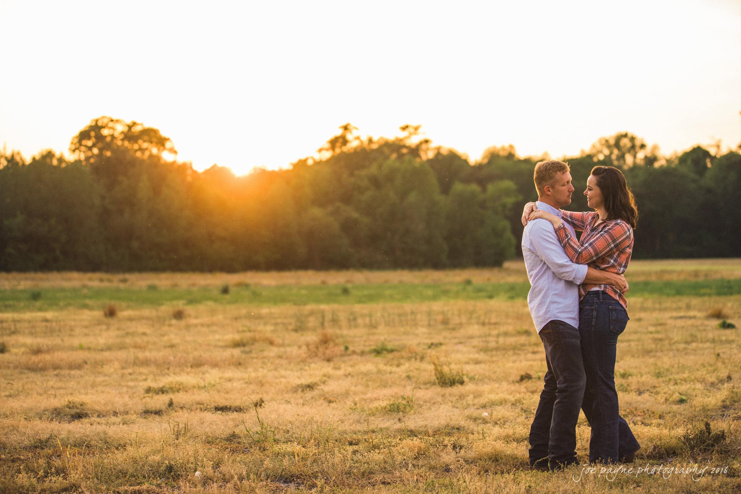 A couple embraces in a field at sunset. They are standing close, gazing into each others eyes, with trees silhouetted against the golden sky in the background.