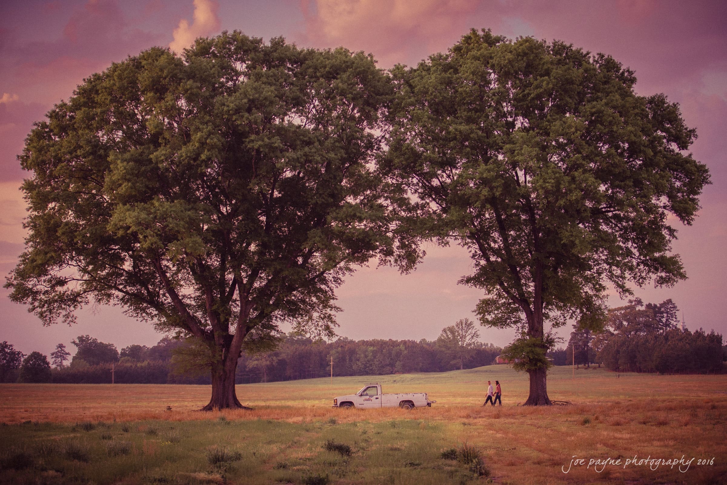 A couple walks hand in hand near a white pickup truck parked between two large trees in a field at sunset. The sky is filled with pink and purple hues, creating a serene and romantic atmosphere.