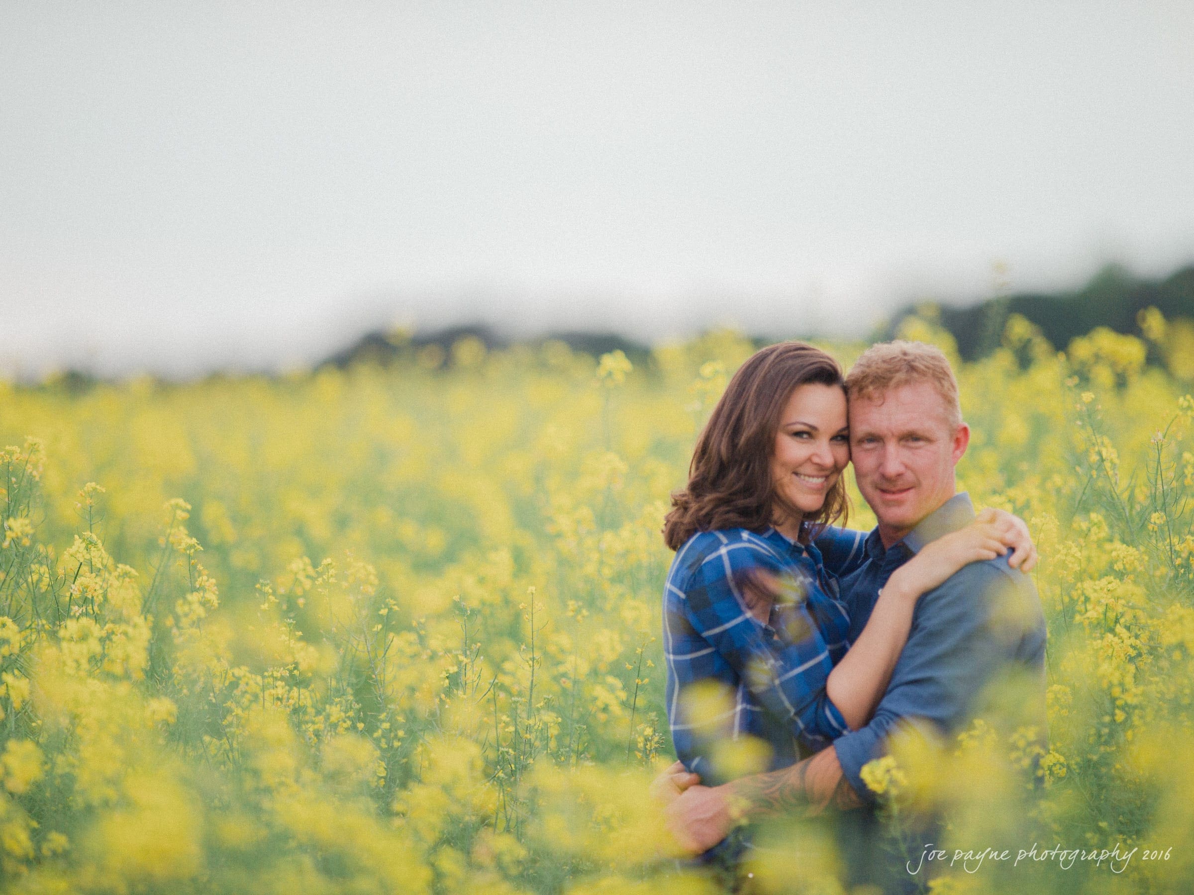 A couple embraces in a field of blooming yellow flowers. The sky is overcast, and the couple is wearing casual clothing. They appear content and relaxed, surrounded by the vibrant blossoms.