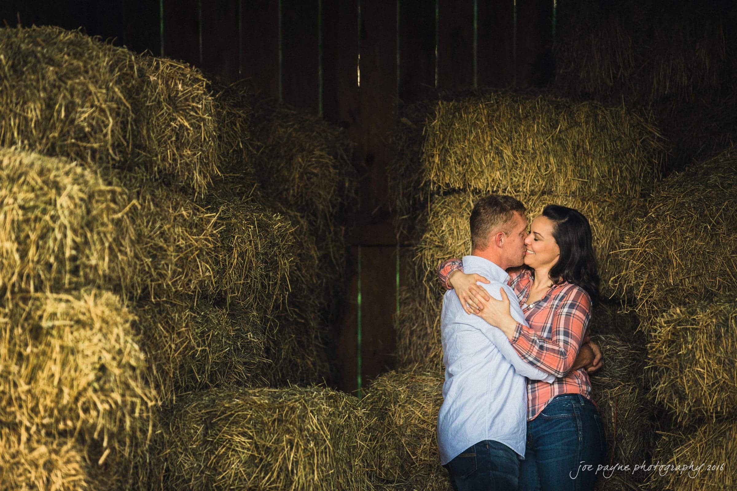 A couple embraces and smiles in a barn surrounded by stacks of hay bales. The man is wearing a light blue shirt, and the woman is wearing a plaid shirt. The scene is warmly lit, creating a cozy and intimate atmosphere.