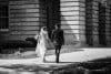 A black and white photo of a bride and groom walking hand in hand on a path. The bride is wearing a long gown and veil, holding a bouquet. The groom is in a suit. They are walking toward a large stone building.