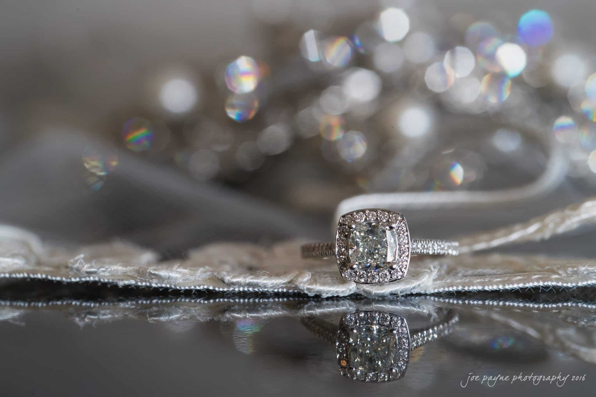 the oaks at salem wedding photography - nicole & brendan A close-up of a diamond engagement ring with a square-cut stone and a halo setting, resting on a lace surface. The background features a soft focus of sparkling lights.