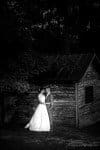 A bride and groom stand closely, sharing an intimate moment in front of a rustic wooden building. The bride wears a flowing white gown, and the groom is in a suit. The setting is dimly lit, adding a romantic, timeless feel.