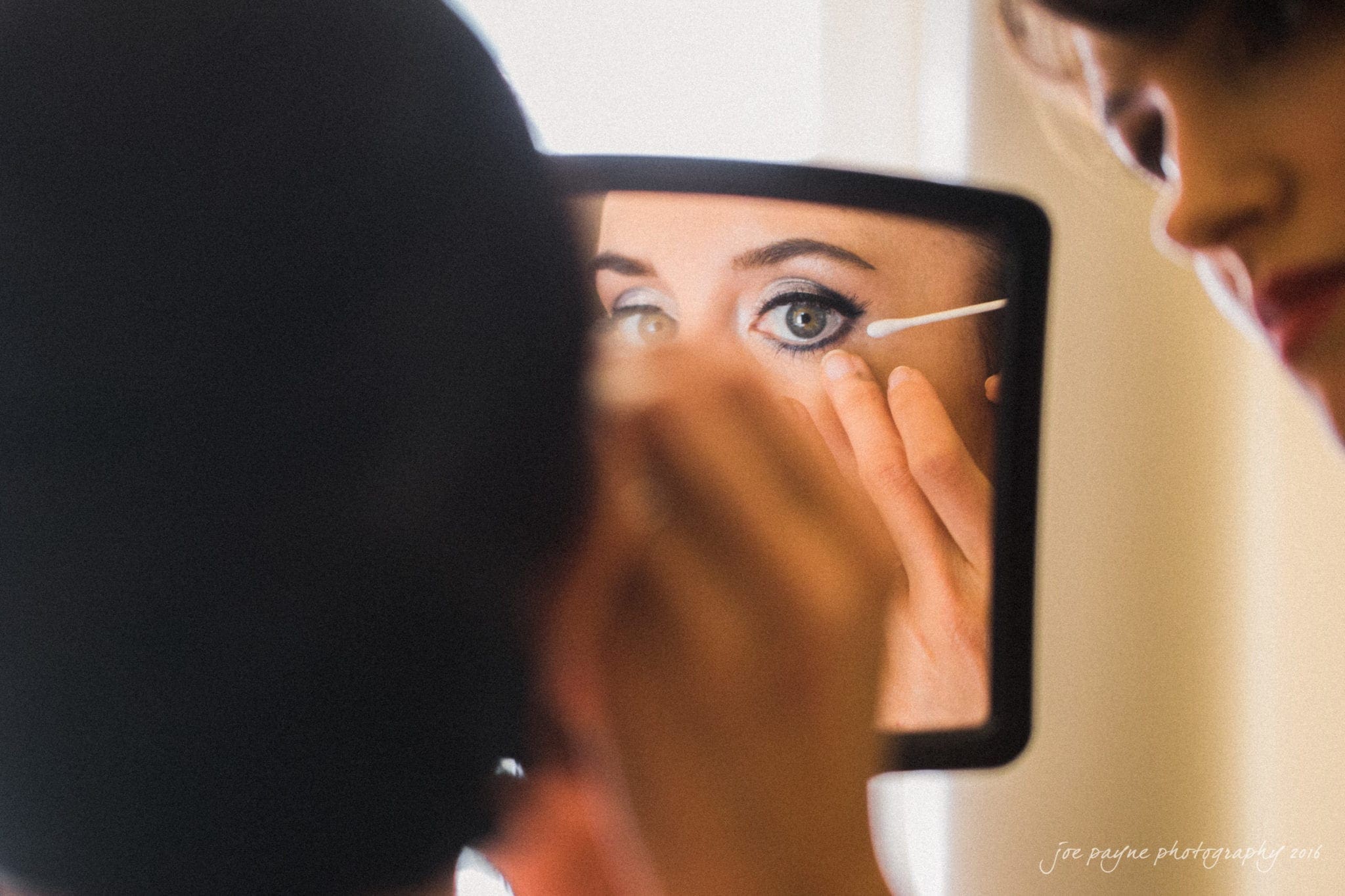 the oaks at salem wedding photography - nicole & brendan A woman holds a small mirror while applying eyeliner. Her eye is focused, and she uses a cotton swab to perfect the makeup under her eye. Another person partially visible watches or assists. The scene is softly lit, highlighting her eye makeup.