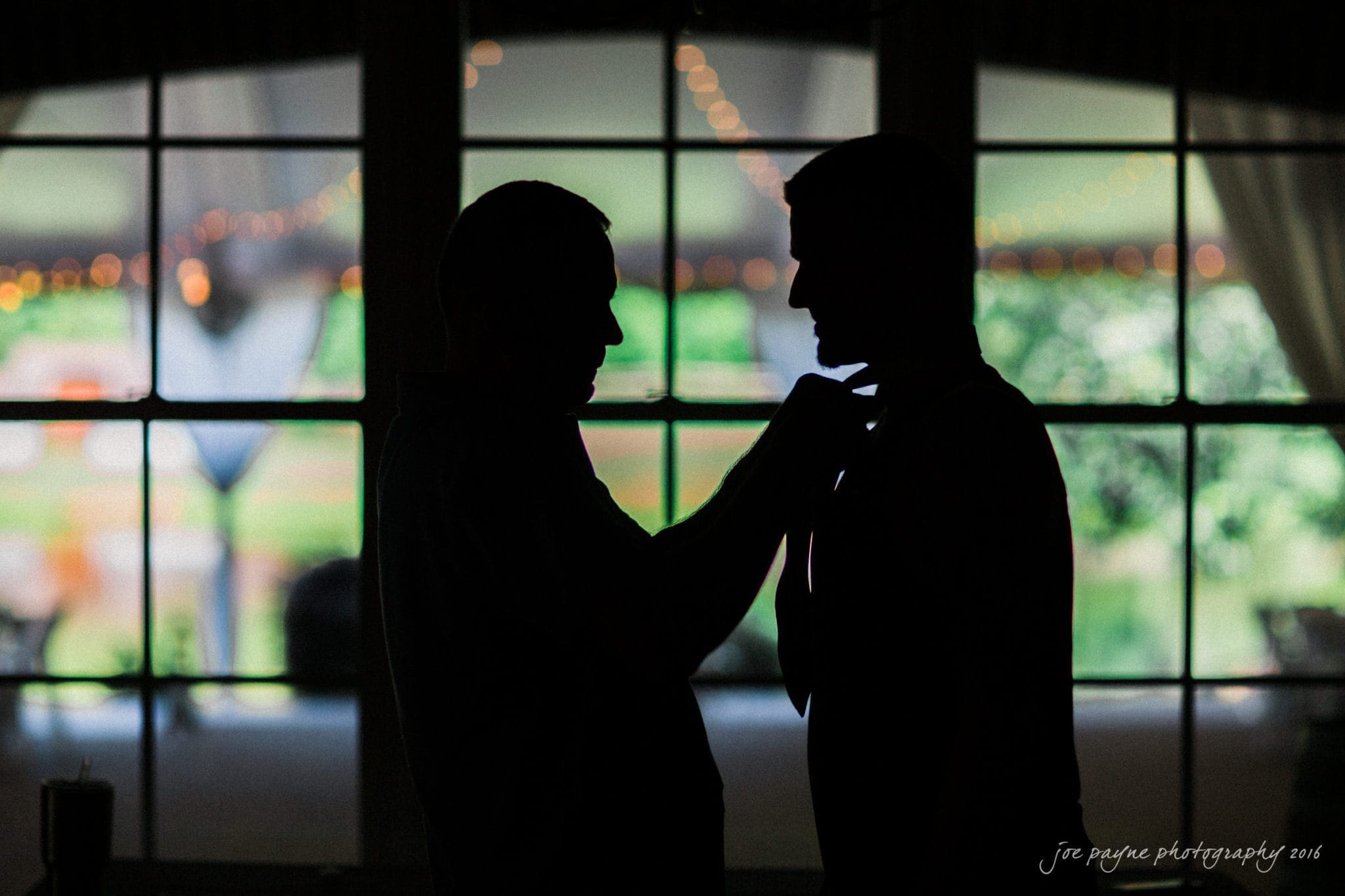 the oaks at salem wedding photography - nicole & brendan Silhouette of two people indoors, one adjusting the others tie in front of a large window. Warm, ambient lights are visible in the background, creating a soft and intimate atmosphere.